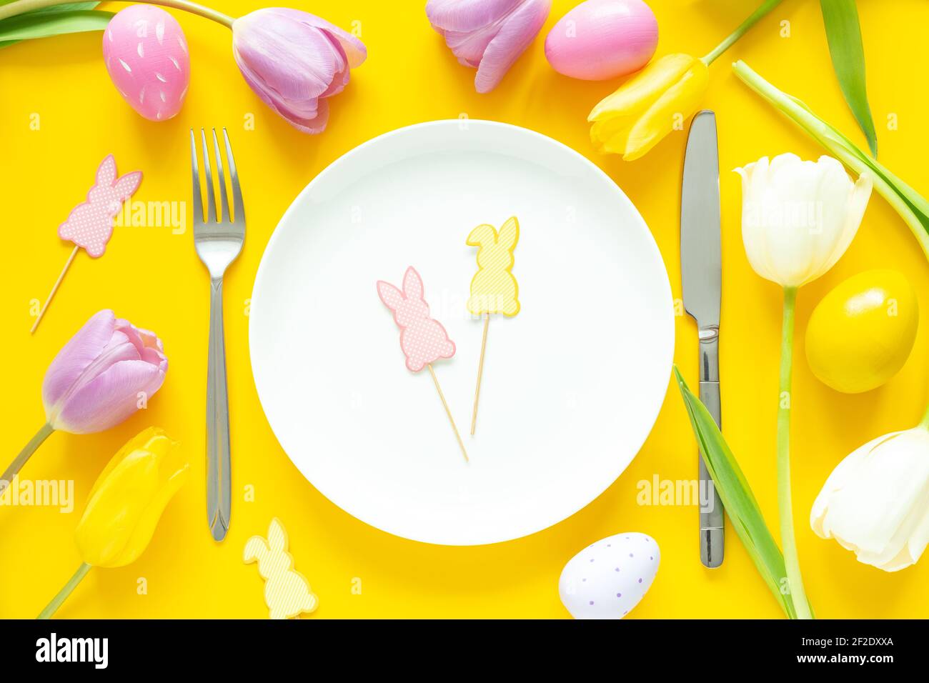 Réglage de la table de Pâques. Assiette blanche, œufs de pâques décoratifs, tulipes et couverts argentés sur fond jaune - image Banque D'Images