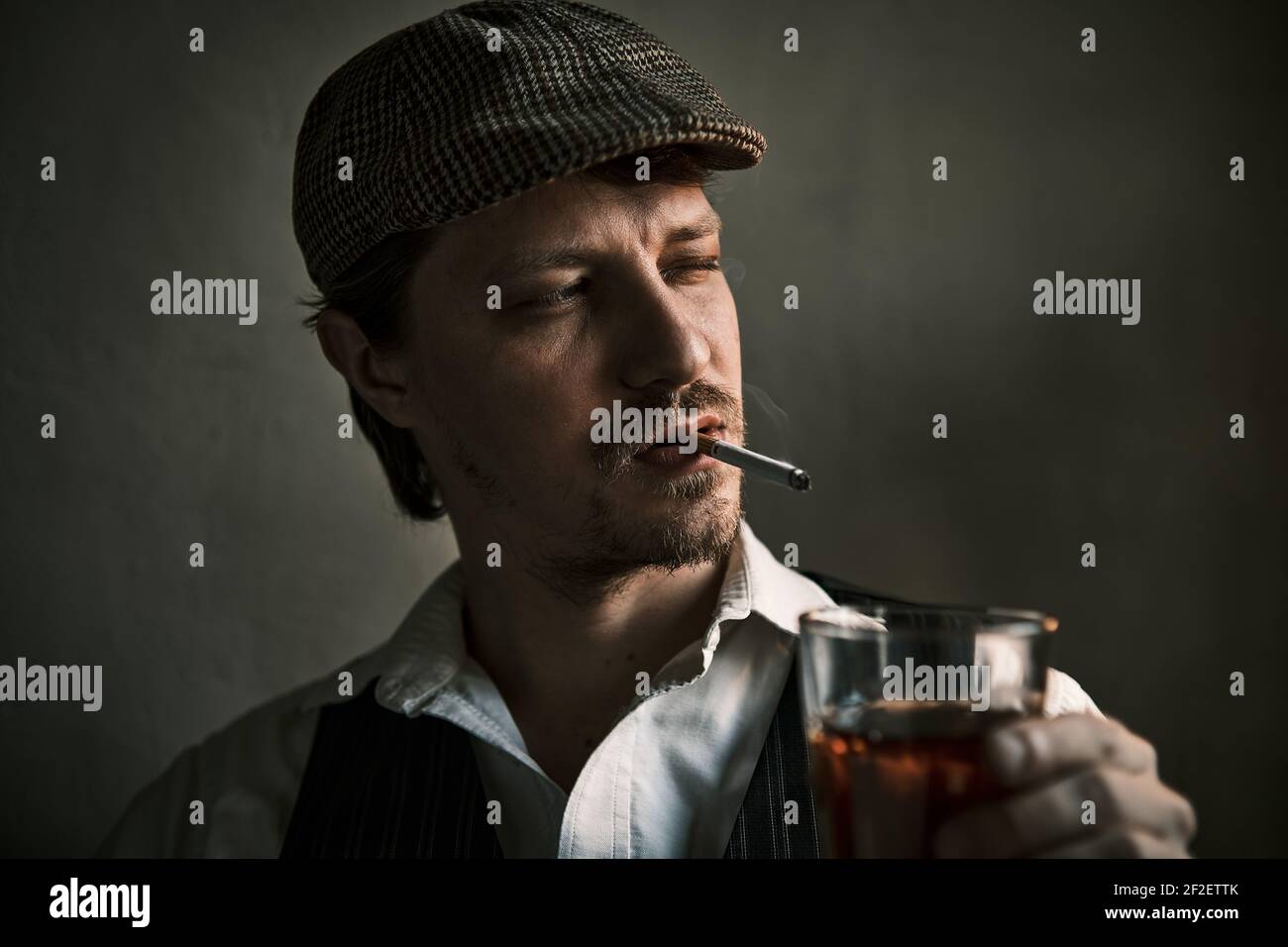 Un jeune homme fume une cigarette et boit du whisky, souffle de la fume,  vtu d'un style rtro, dans un bret, une prise de vue cinmatographique,  un gros plan Photo Stock -