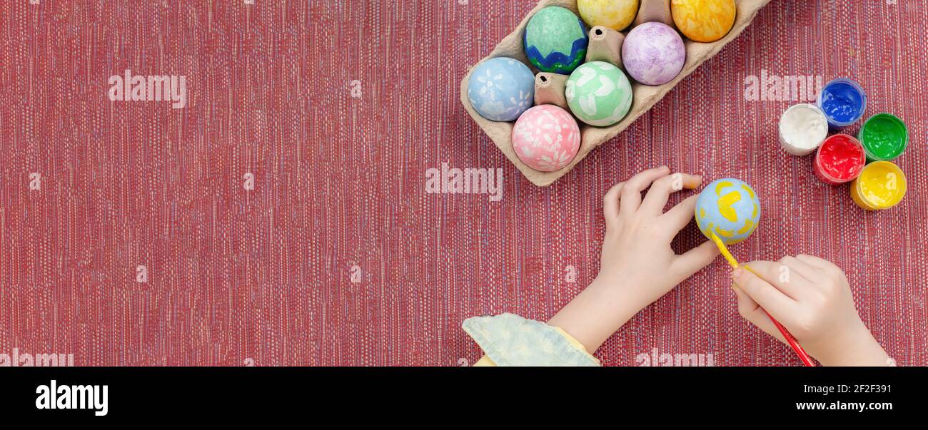 Un garçon peint un œuf de Pâques, vue du dessus sur un fond en tissu rouge. Peinture des oeufs de Pâques, peintures de gouache, pinceau, préparation pour les vacances de Pâques. BA Banque D'Images