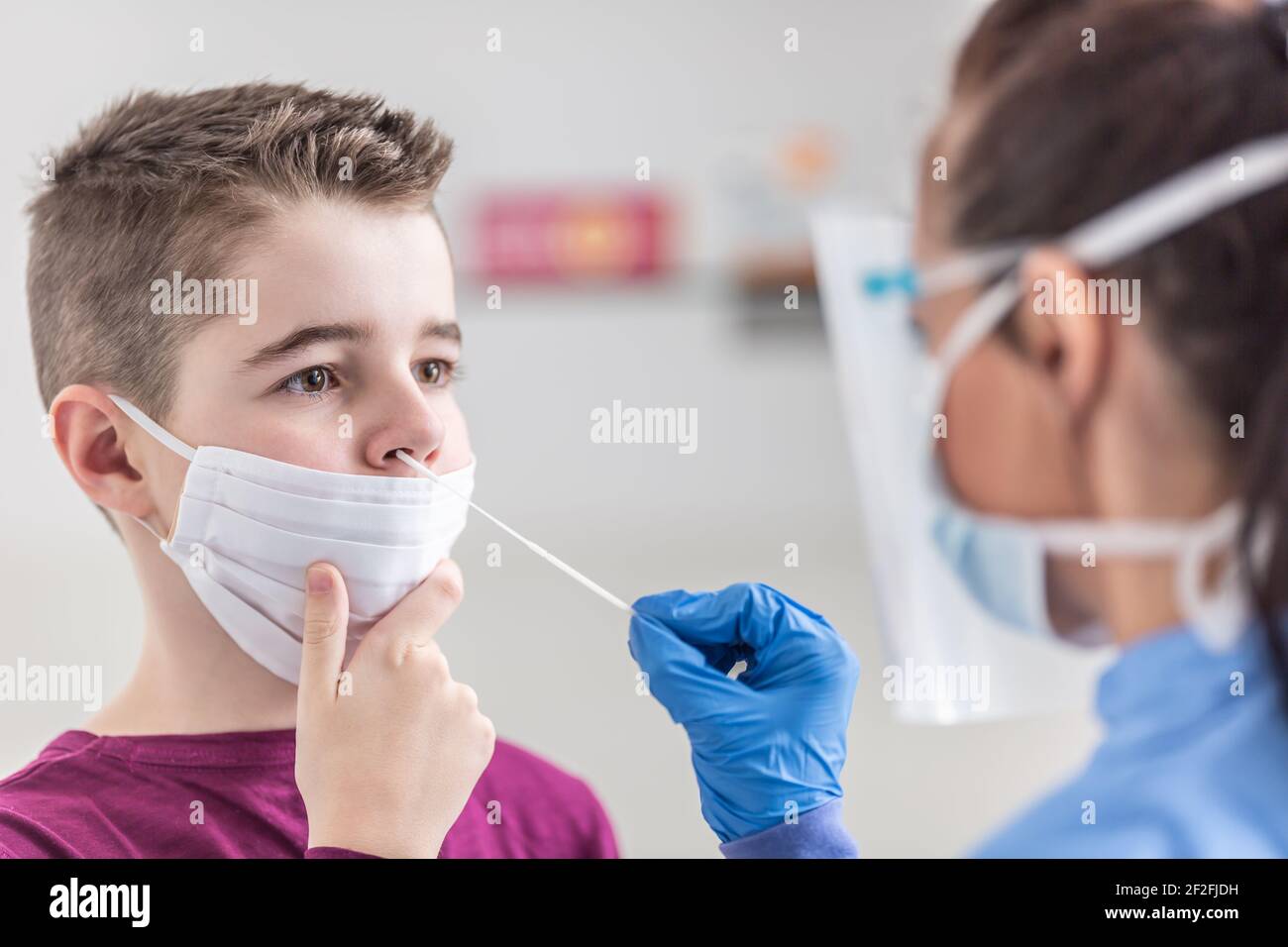 Un jeune garçon portant un masque facial sur la bouche tient encore quand l'infirmière prend un échantillon de son nez à l'aide d'un écouvillon. Banque D'Images