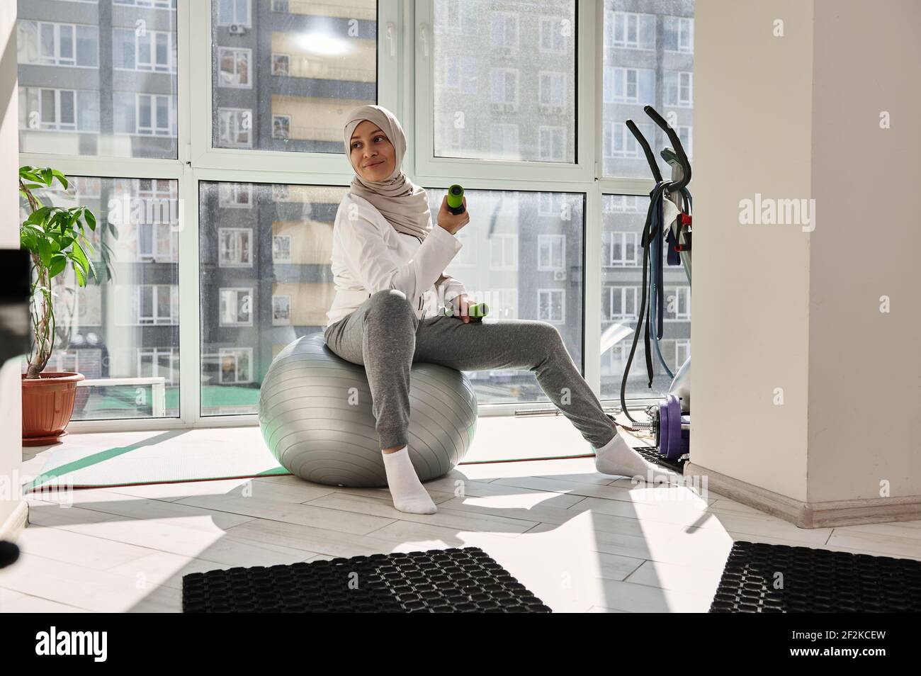 Une jeune fille musulmane souriante s'entraînant dans le hijab avec des haltères à la maison, regardant loin tout en étant assise sur une balle de sport. Entraînement à domicile Banque D'Images