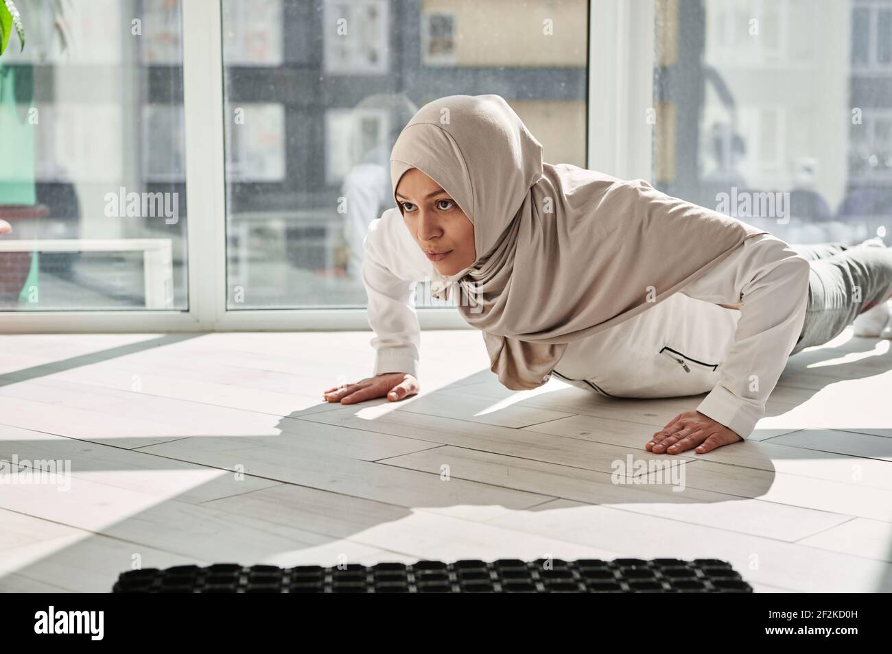 Femme arabe musulmane déterminée faisant des push-up contre le fond de grandes fenêtres à la maison lors d'une belle journée ensoleillée. Concept d'entraînement à domicile Banque D'Images