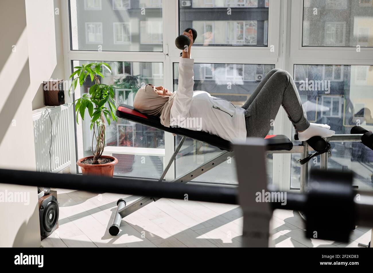 Vue latérale d'une femme arabe musulmane déterminée avec la tête couverte dans hijab faisant dumbbbell banc de presse à la salle de gym à la maison contre l'arrière-plan des grandes fenêtres Banque D'Images