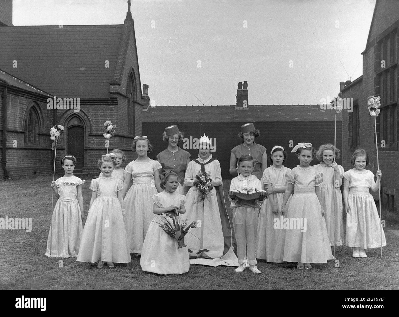1956, historique, dans les jardins de leur église locale, un groupe d'enfants dans leurs jolis grenouille ou robes, de la ligne pour une photo avant le traditionnel défilé ou carnaval de mai, Leeds, Angleterre, Royaume-Uni. La Reine de mai se tient au milieu du groupe, avec un petit garçon tenant un coussin, qui tenait sa couronne. Plusieurs des filles tiennent des bâtons de fleur. Les célébrations du jour de mai sont un festival traditionnel, marquant le début de l'été, datant de l'époque romaine, et le festival de la flore. Banque D'Images