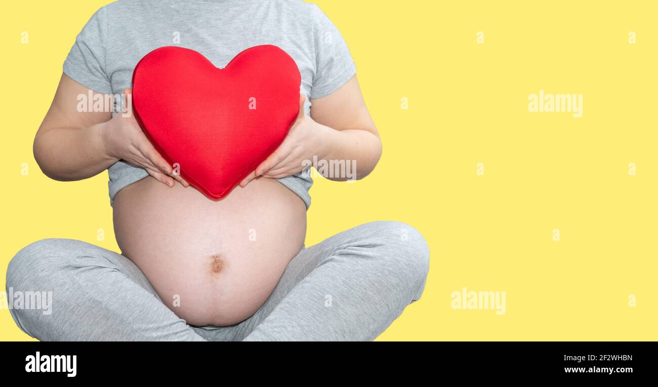 Grossesse, maternité, personnes, amour et attente concept - gros plan d'une femme enceinte tenant un coussin en forme de cœur rouge sur bac jaune illuminant Banque D'Images