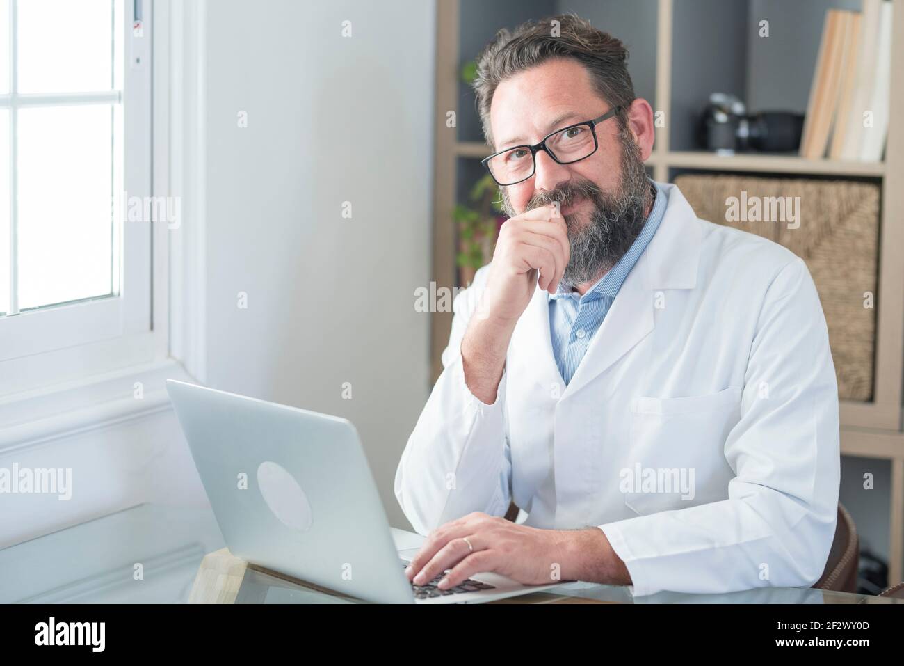 Un vieux médecin souriant en lunettes et un uniforme blanc s'assoir à l'hôpital sur un ordinateur portable écrire dans le journal, un homme mature heureux médecin de premier plan remplir p Banque D'Images
