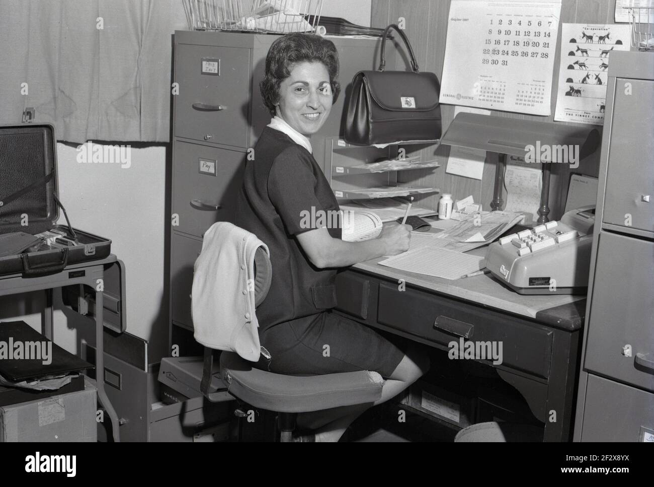 1967, historique, une dame gaie compte personne assise sur une chaise pivotante à un petit bureau contre un mur, avec ses papiers et une grande calculatrice mécanique. Divers plateaux de caisse, un calendrier au mur et deux armoires de rangement en métal profond se tiennent de chaque côté du bureau en bois dans le petit bureau, un peu exigu. Banque D'Images