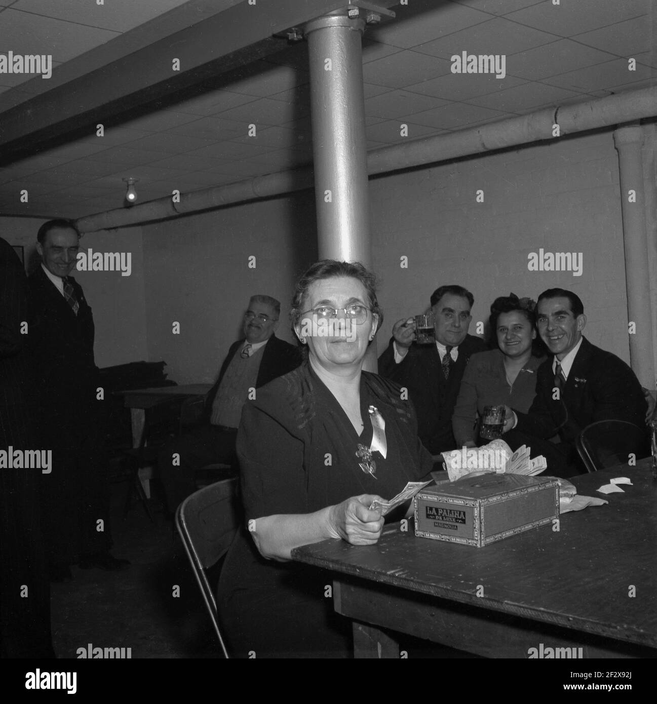 Fin des années 1940, historique, près de la salle d'horloge dans une cave ou un sous-sol clalub ou un bar, une femme mature caissière assis à une table avec une boîte à cigares « la Palina de Luxe » comptant des billets de dollars, comme les clients derrière elle aiment un verre et un rire. Une marque célèbre de cigares américains fabriqués par la Congress Cigar Company et dirigée par Sam Paley, père du fondateur de CBS, William S. Paley, la marque a été créée à Chicago au milieu des années 1800. La Palina est un dérivé de 'la femelle Paley et l'épouse de Sam Paley, Goldie Drell Paley est apparu dans le costume espagnol sur la boîte. Banque D'Images
