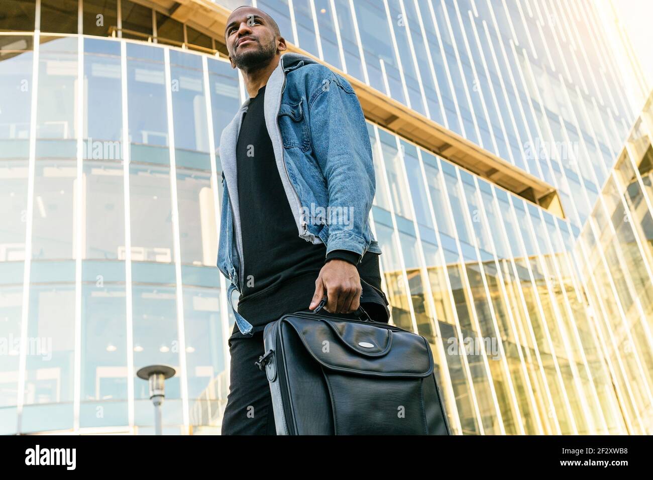 Vue d'en dessous de côté de l'homme ethnique adulte dans une ambiance décontractée tenue avec porte-documents en cuir se promenant en ville tout en regardant loin Banque D'Images