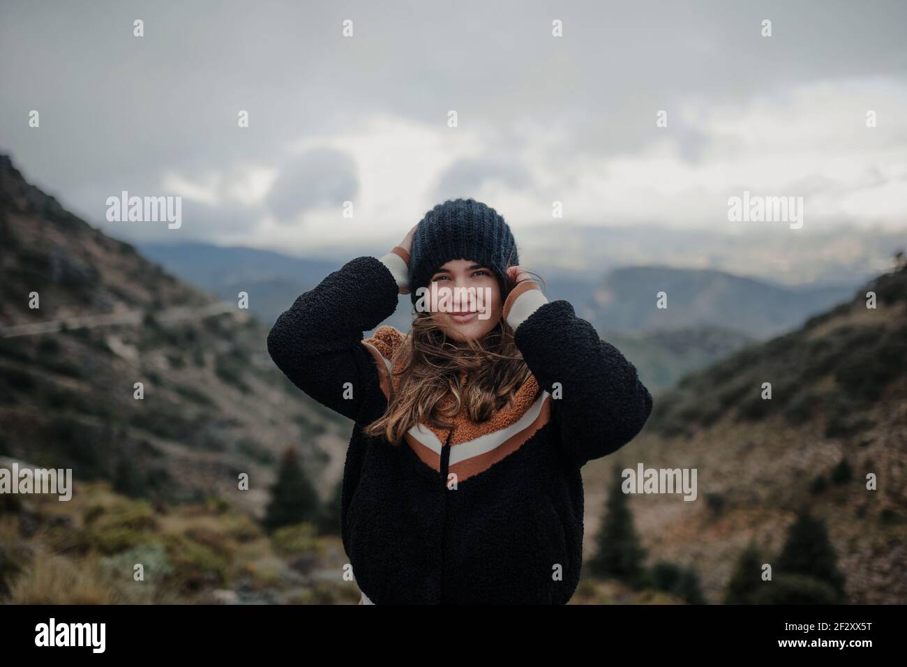 Jeune femme souriante qui met un chapeau chaud et regarde caméra en position debout sur des hauts plateaux accidentés par un jour sombre et nuageux Banque D'Images