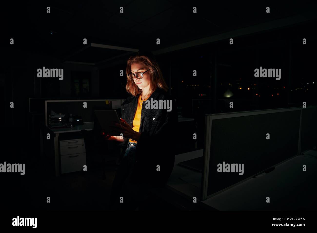 Jeune femme d'affaires créative portant des lunettes sur une tablette numérique tard à nuit dans le bureau sombre Banque D'Images