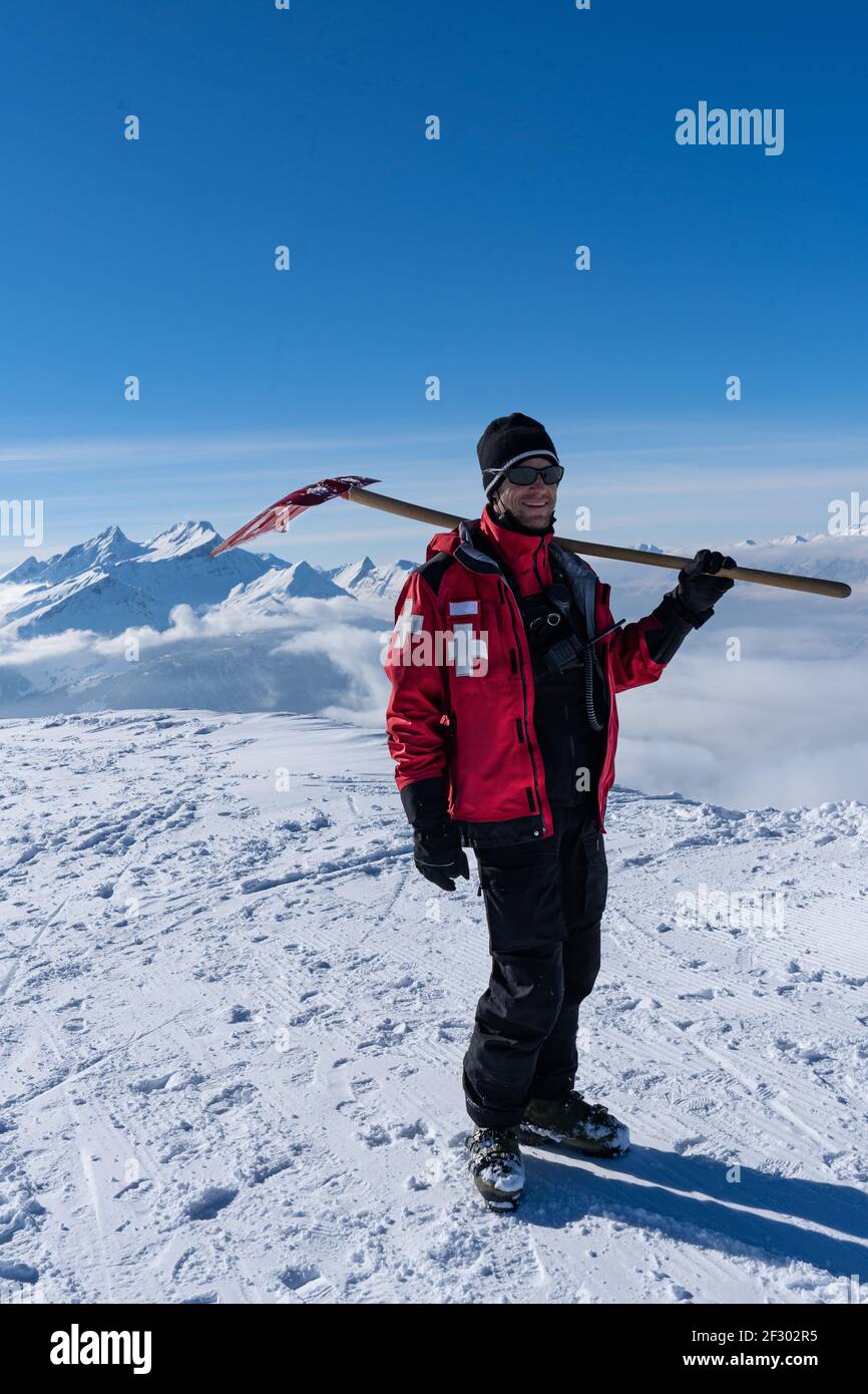 ski patroller sur montagne enneigée est debout contre le soleil avec une veste de sauvetage rouge et une pelle sur ses épaules. Un homme vérifie le mât gelé o Banque D'Images