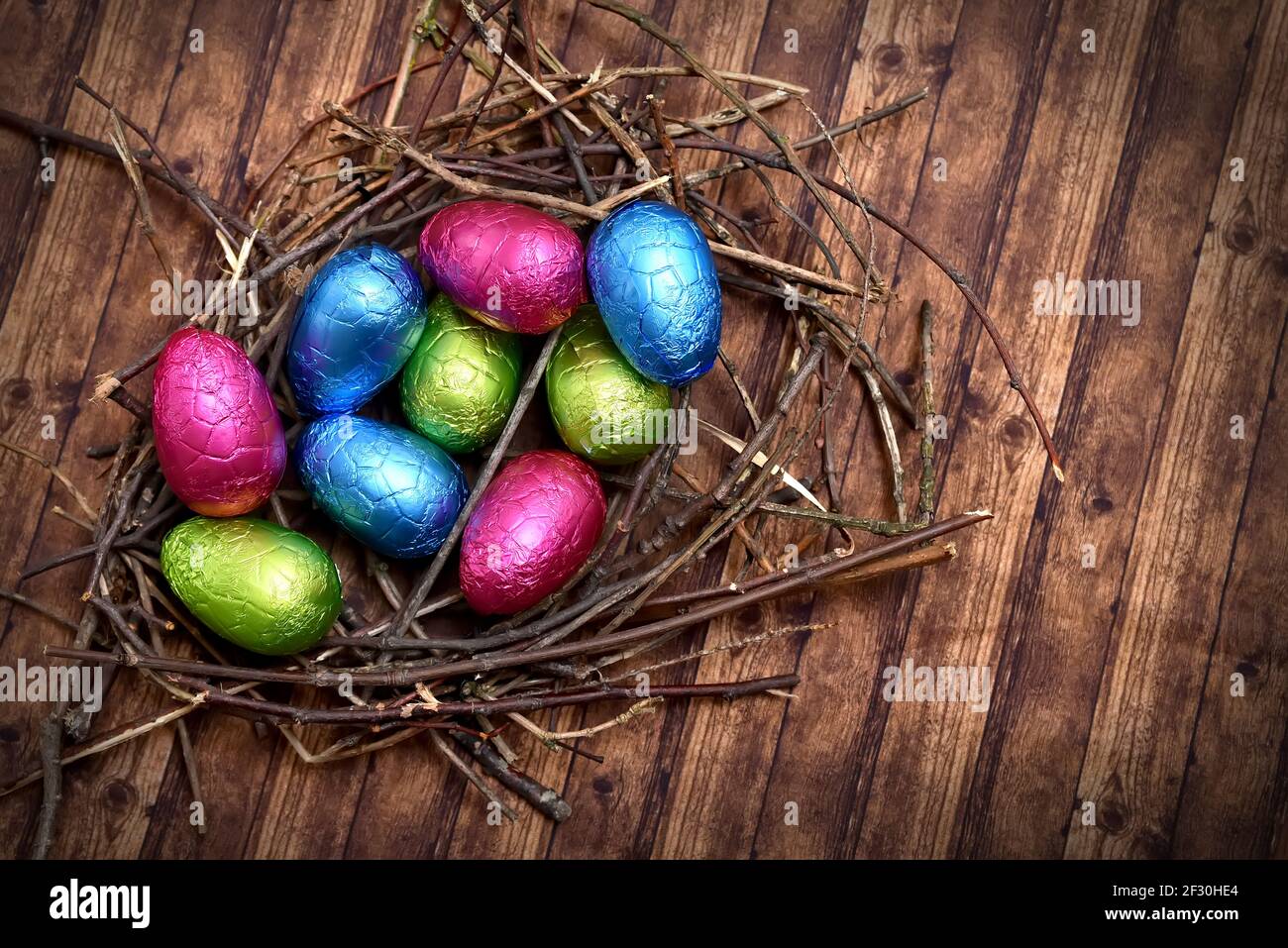 Des œufs de pâques colorés enveloppés de feuilles dans du rose, du vert, du bleu et du jaune dans un nid naturel fait de bâtons et de brindilles, contre un dos en bois brun multigrain Banque D'Images