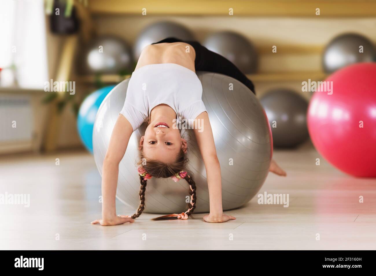 Une petite fille de vêtements de sport fait un exercice de pont sur un fitball dans un studio de sport. L'enfant sourit et pose avec sa tête vers le bas, en inclinant ses bras f Banque D'Images