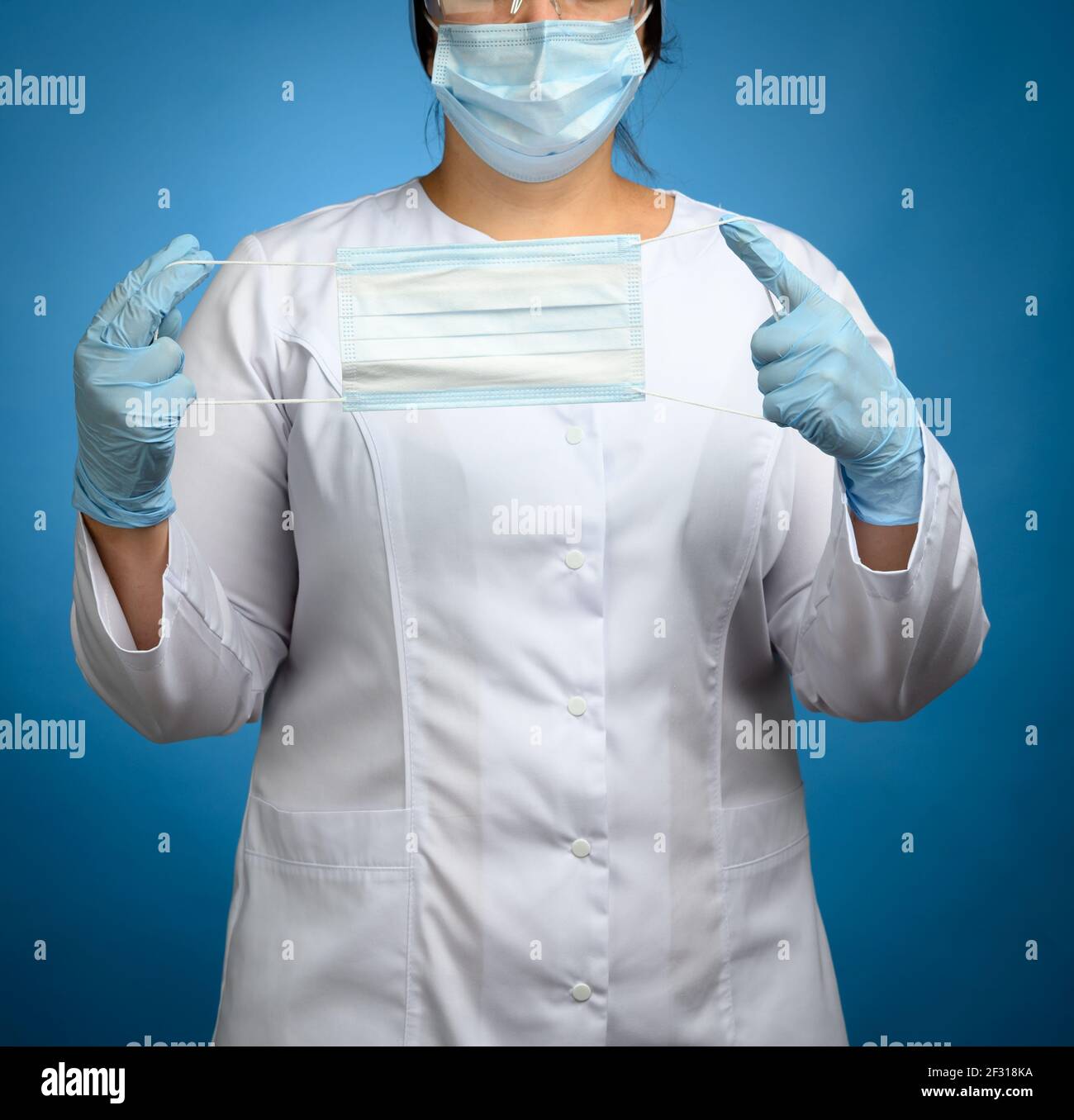 Femme médecin dans un manteau médical blanc, un masque jetable, des lunettes de protection en plastique et une casquette se tient sur un fond bleu et ho Banque D'Images
