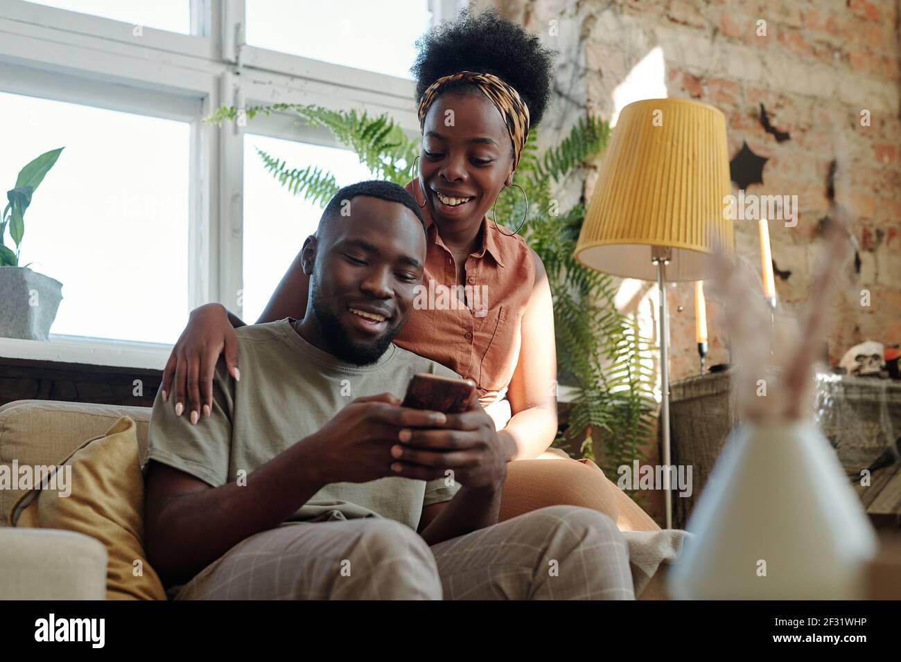 Joyeux jeune couple africain en vêtements décontractés regardant l'écran de smartphone tenu par un homme souriant assis devant caméra contre fenêtre Banque D'Images