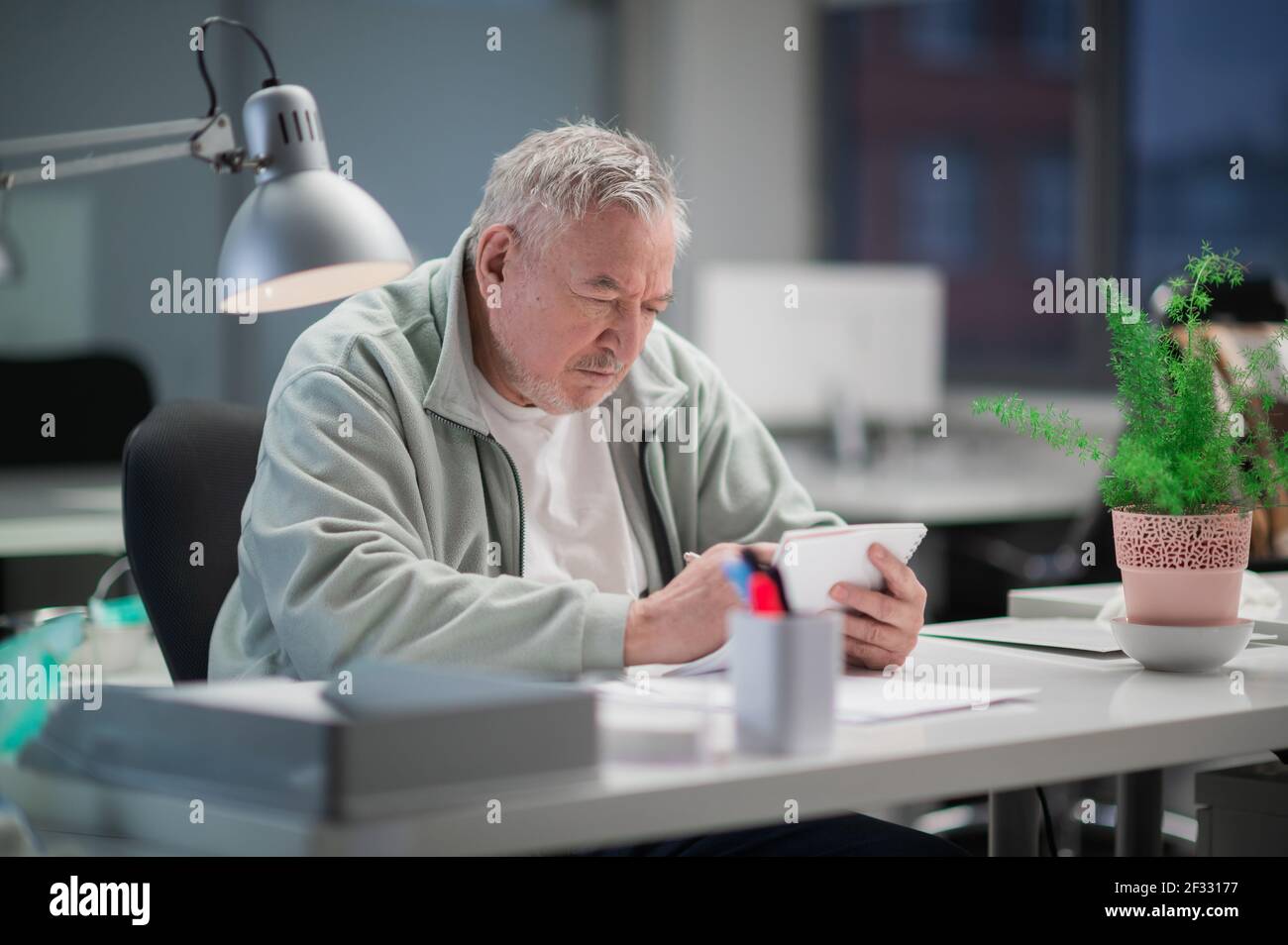 Un homme de bureau âgé aux cheveux gris lit un texte dans un bloc-notes. Un grand bureau lumineux avec des plantes et un ordinateur en arrière-plan Banque D'Images