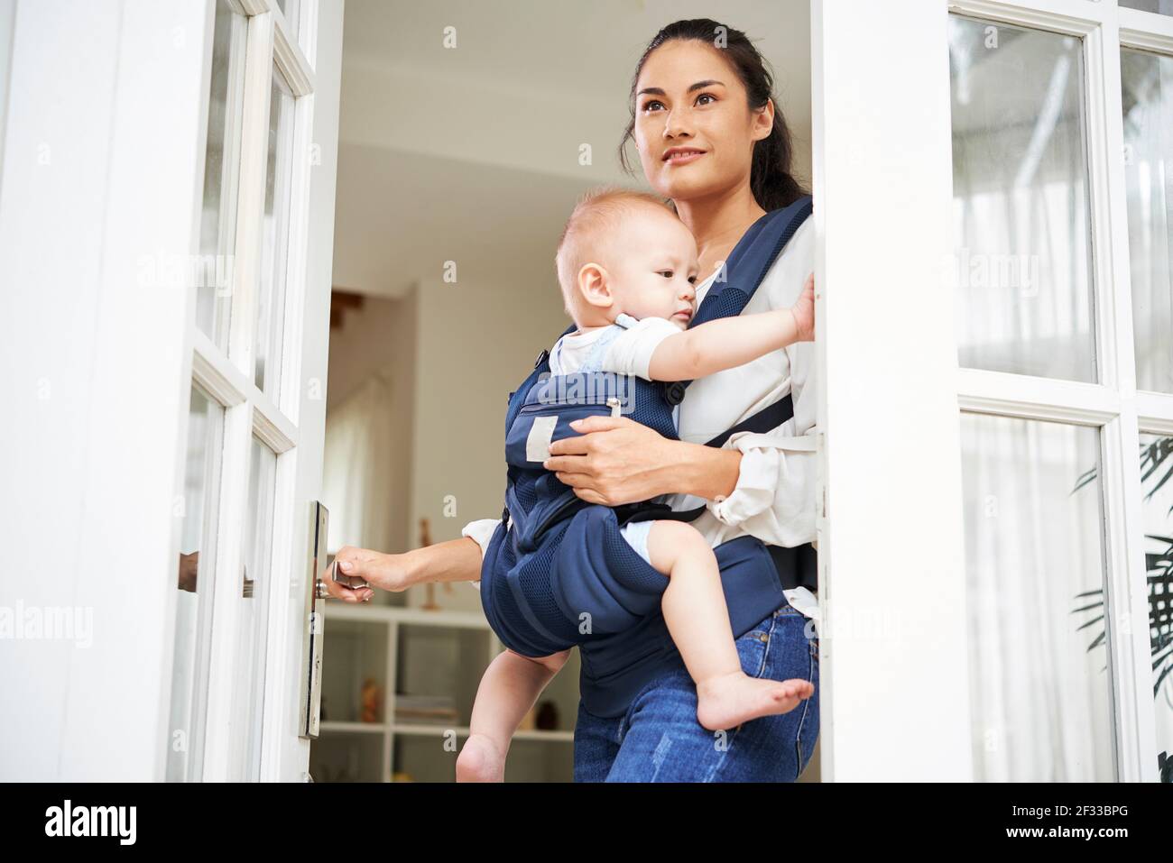 Jolie jeune femme de course mixte ouvrant la porte d'entrée et quittant la maison avec bébé garçon dans le support Banque D'Images