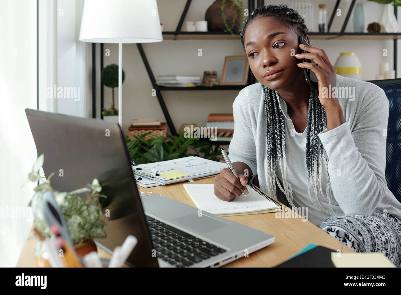 Jeune femme noire sérieuse dans un salon parlant au téléphone avec un collègue, lisant un document sur l'écran d'un ordinateur portable et prenant des notes Banque D'Images