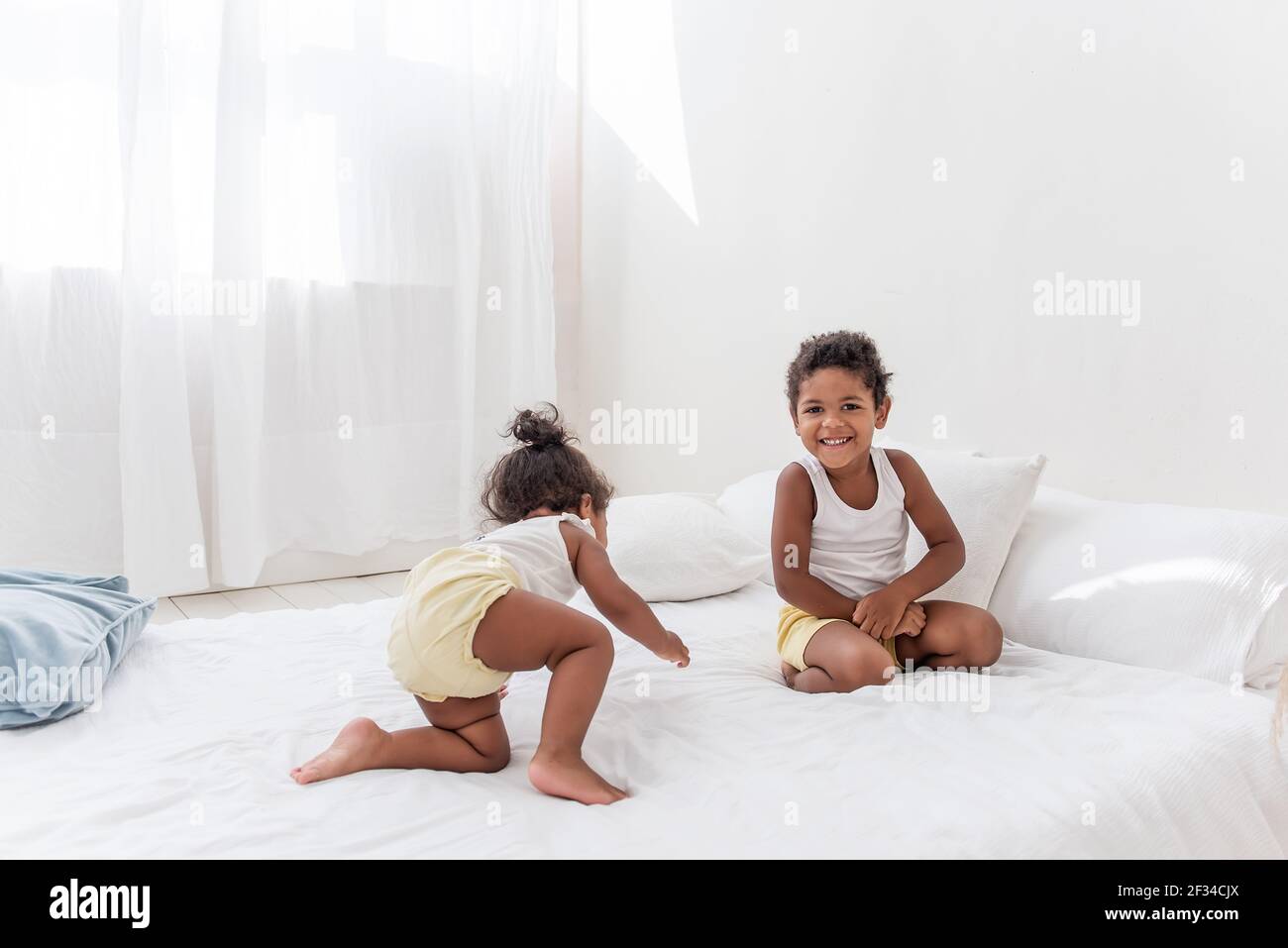 Frère et soeur Afro-Américains jouent ensemble sur un lit blanc dans un loft intérieur. Les frères et sœurs s'amusent parmi les oreillers bleus le matin. Pul. Garçon Banque D'Images
