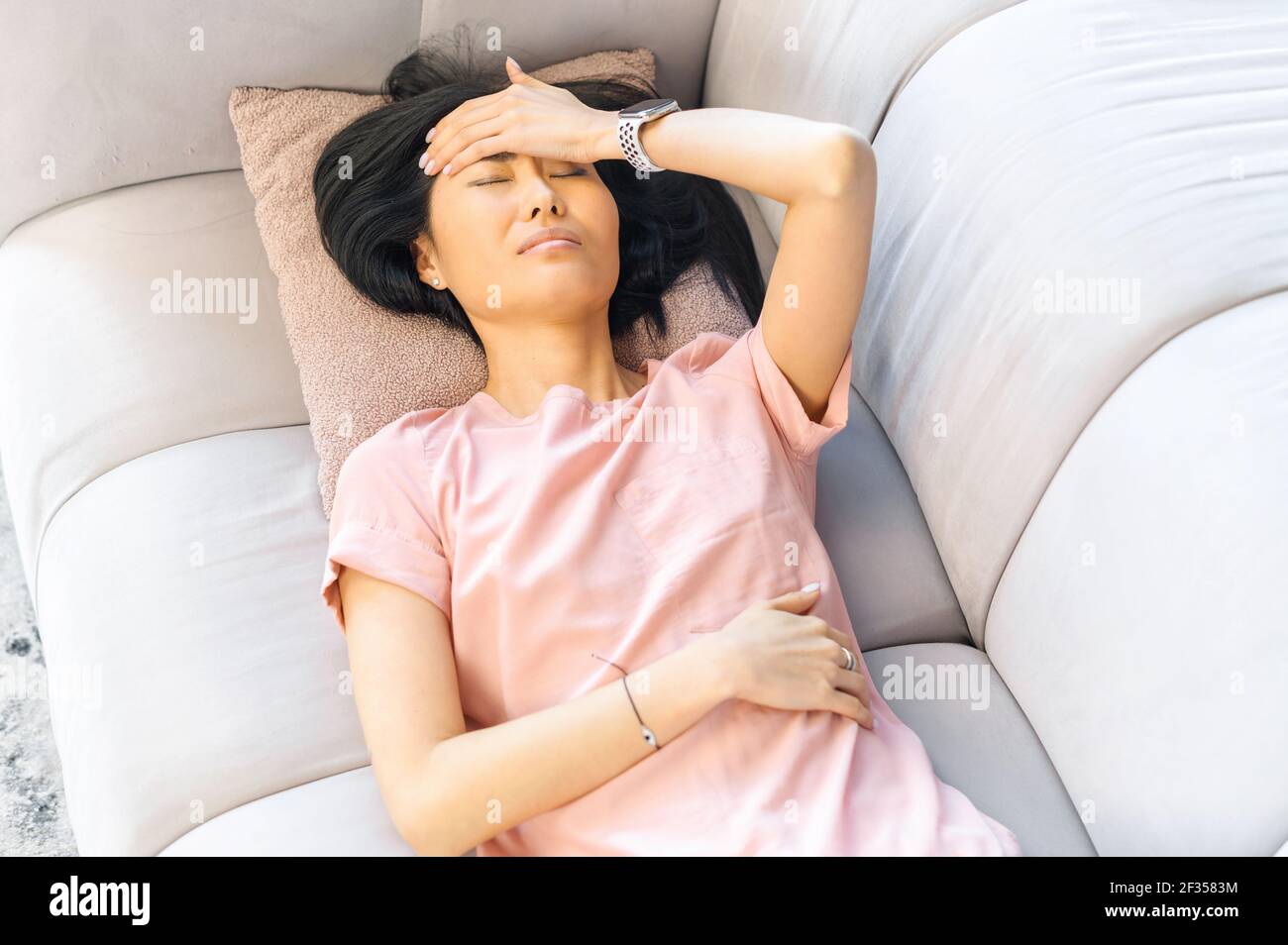 Femme asiatique avec cheveux noirs ayant un mal de tête couché sur le  canapé gris, malade à la maison, avec des douleurs d'estomac, sensible aux  intempéries, malaise, pendant la menstruation, tenez-vous à