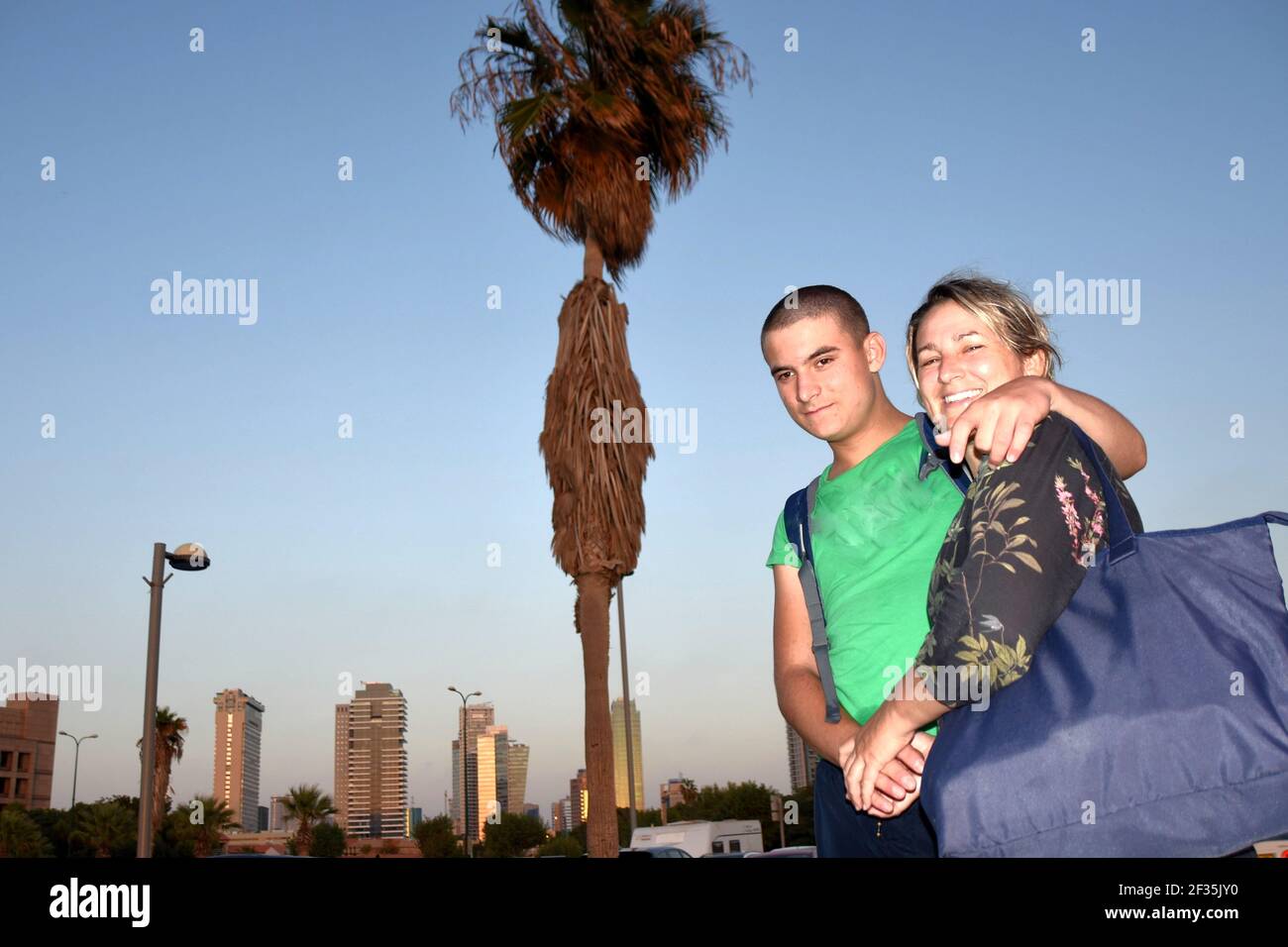 Mère et fils en vacances de retour de la plage dans la ville de tel Aviv en début de soirée souriant à la caméra. Il y a des gratte-ciel et un palmier. Banque D'Images