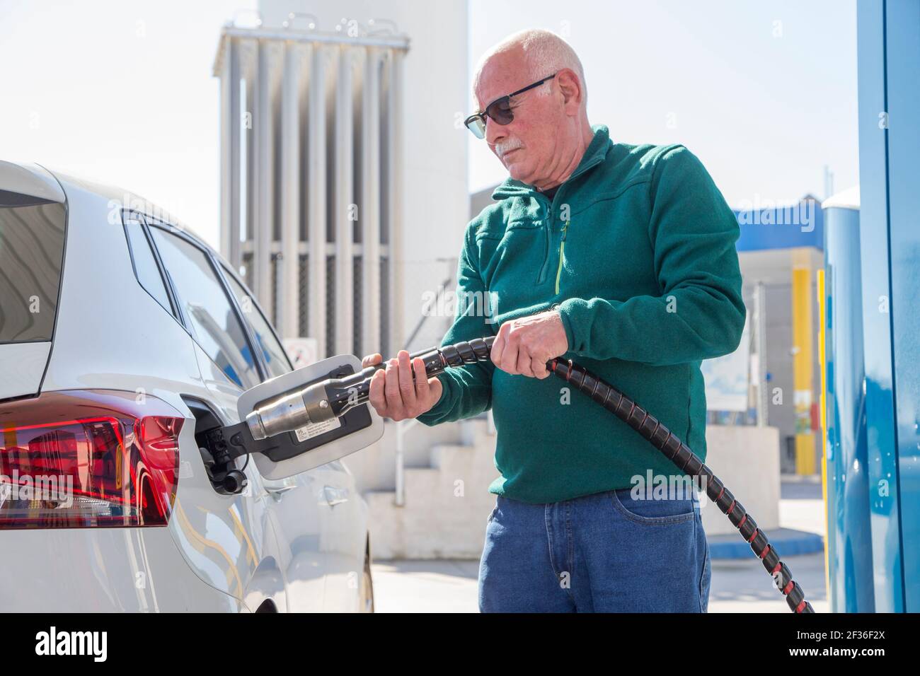 Vieil homme avec chandail vert et jeans tenant le tuyau de gaz naturel comprimé (GNC) pour remplir le réservoir de son éco-voiture. Avec le réservoir de gaz au niveau du backgroun Banque D'Images