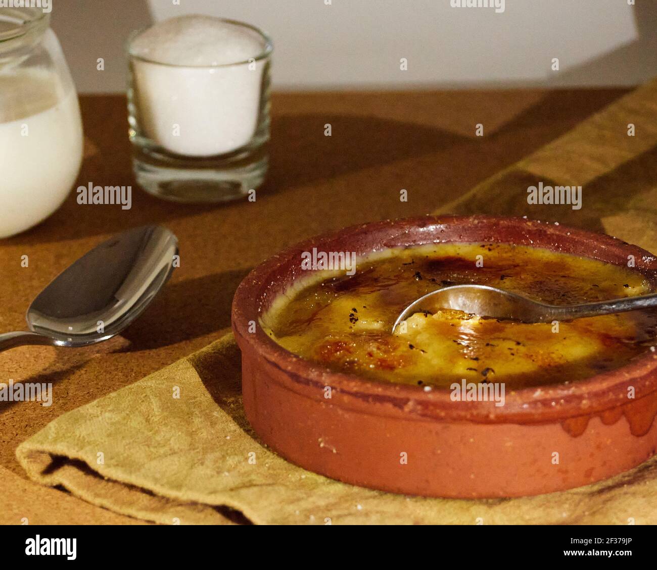 Cuillère plongée dans la crème catalane sur une serviette décorée d'une tasse de sucre et d'une tasse de lait. Sur la table en bois Banque D'Images