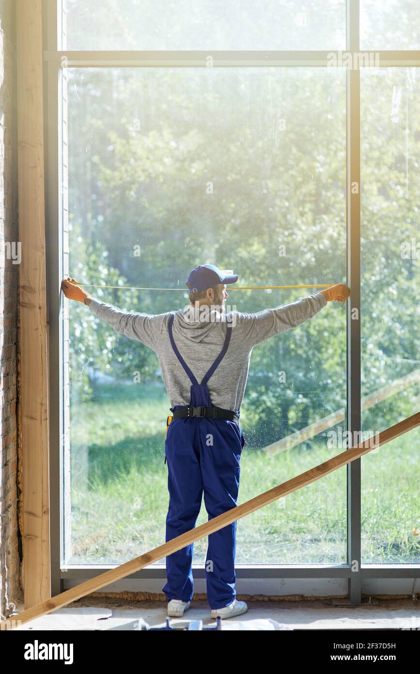Prise de vue en longueur d'un jeune ouvrier portant une combinaison bleue avec mesurer le ruban pendant les travaux sur le chantier de construction de chalets Banque D'Images