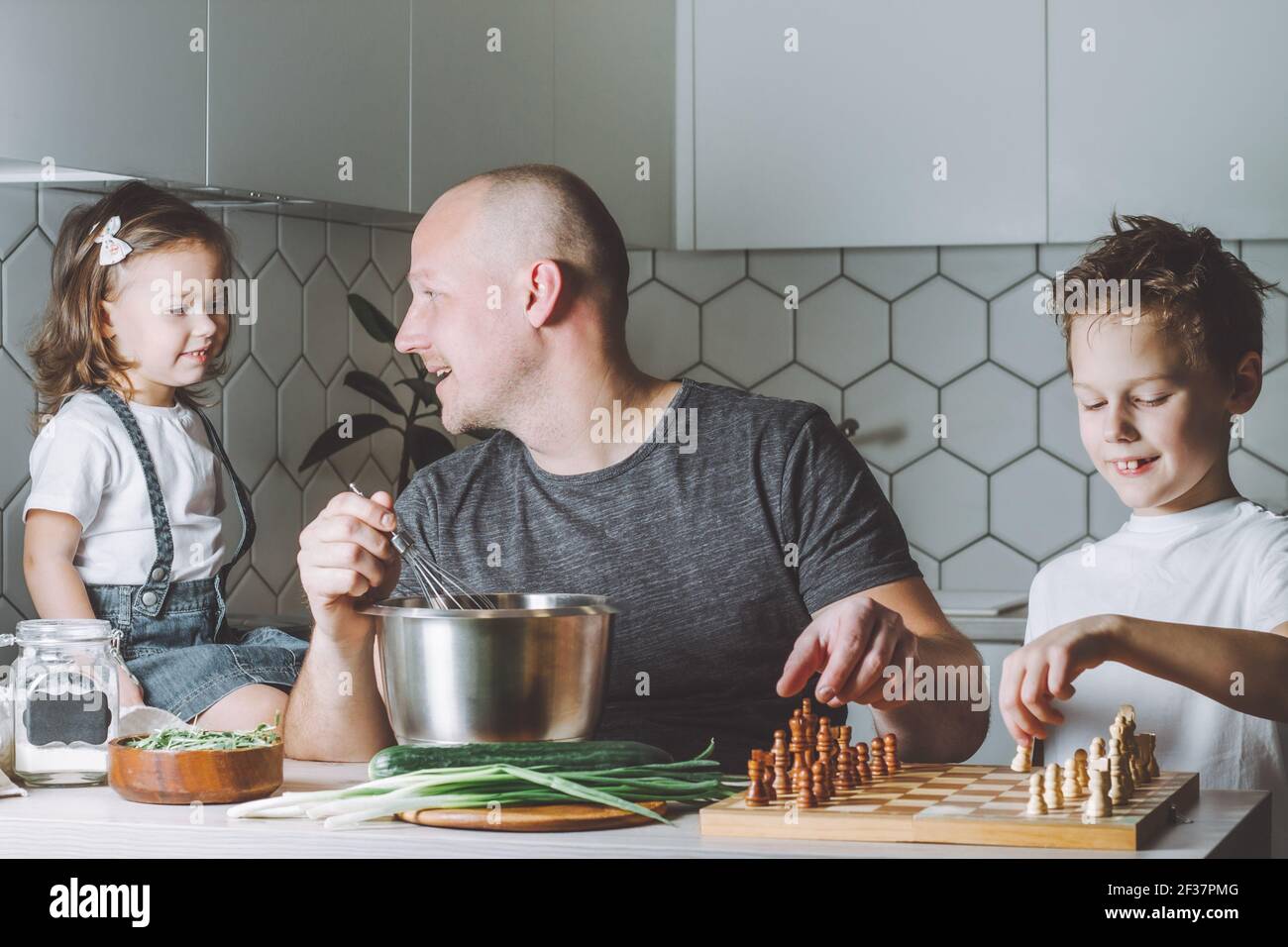 Le père fouette une omelette avec un fouet, jouant aux échecs avec son fils et parlant à sa fille. Homme faisant des tâches ménagères. Banque D'Images