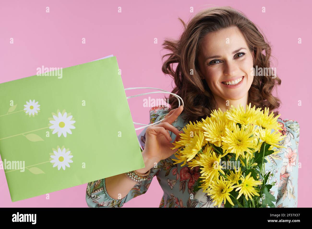 Portrait d'une femme moderne souriante avec de longs cheveux de brunette ondulés avec des fleurs de chrysanthèmes jaunes et un sac de shopping vert sur fond rose. Banque D'Images