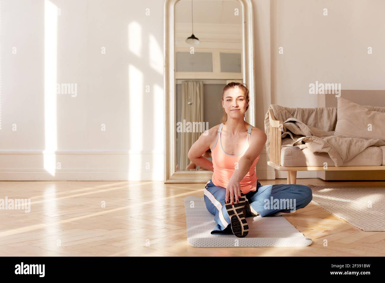 Une athlète féminine souriante dans des vêtements de sport assis sur un tapis dans la vie faites de la place et étirez vos jambes tout en vous réchauffant avant de vous entraîner à la maison et regarder l'appareil photo Banque D'Images