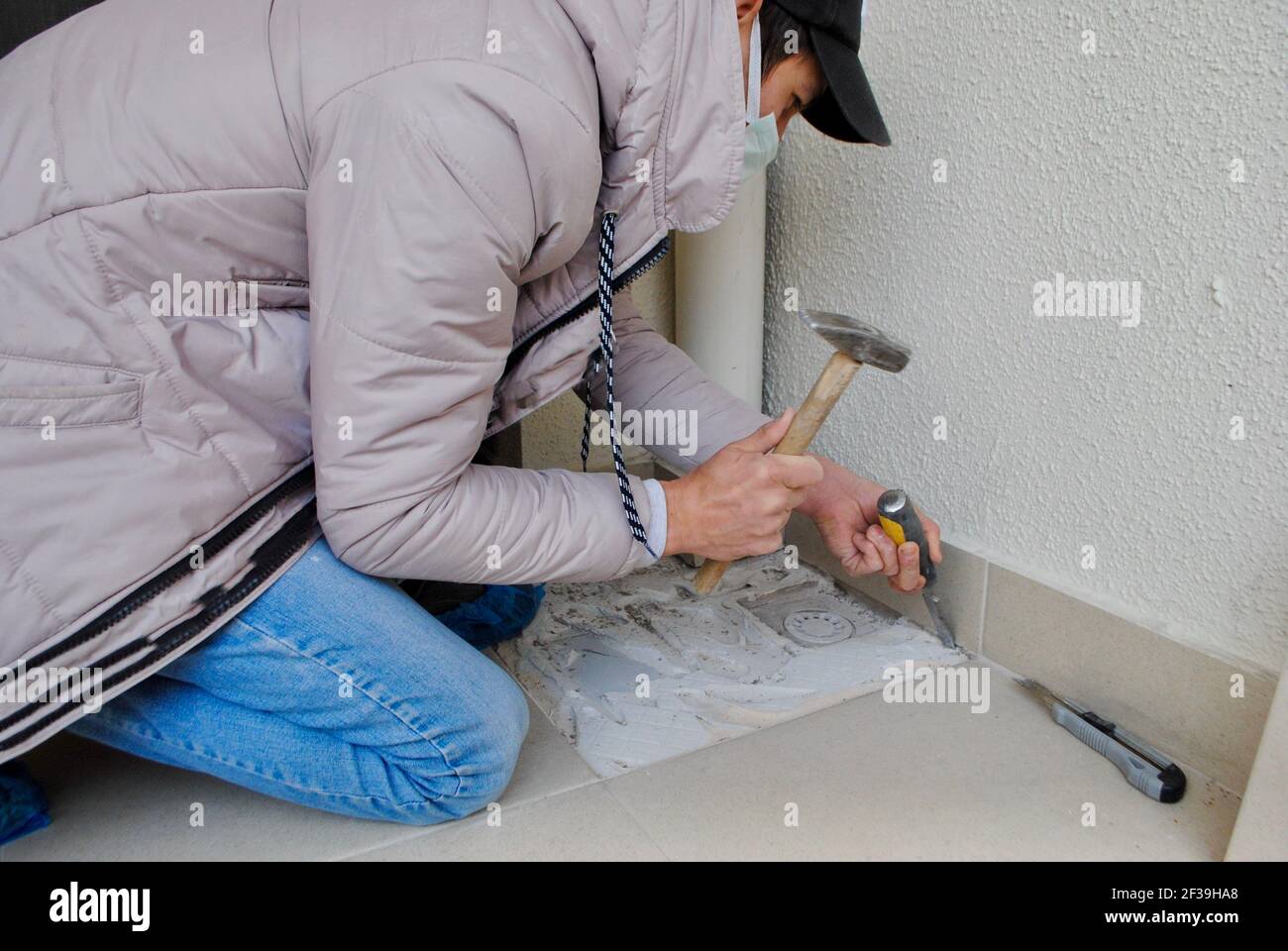 Homme avec masque enlève une tuile cassée dans le balcon avec un marteau et un burin. Démontage de l'ancien plâtre et de la colle de ciment pour un nouveau carreau. Rénovation. Accueil Banque D'Images