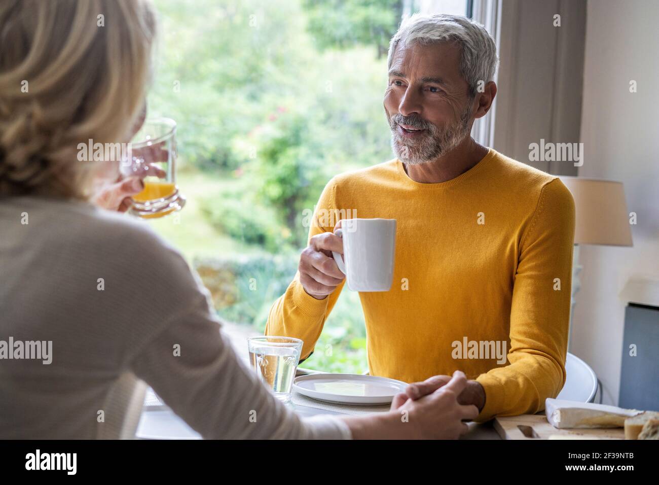 Couple mûr souriant tenant les mains tout en prenant le petit déjeuner à la maison Banque D'Images