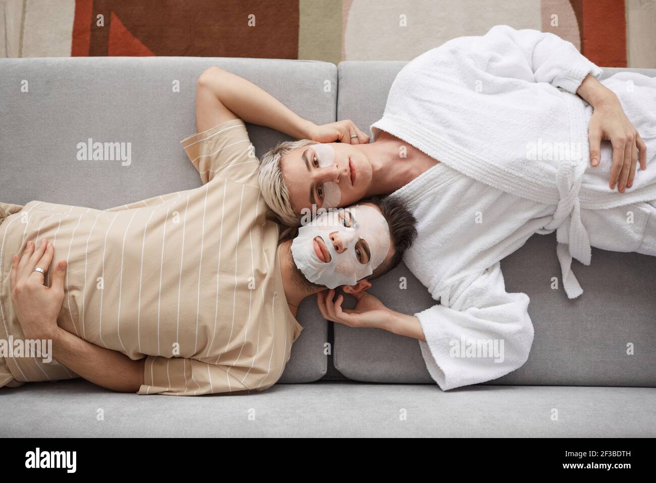 Vue de dessus portrait d'un jeune couple gay regardant la caméra tout en étant allongé sur un canapé ensemble et portant des masques faciaux, beauté et concept de soin de la peau Banque D'Images