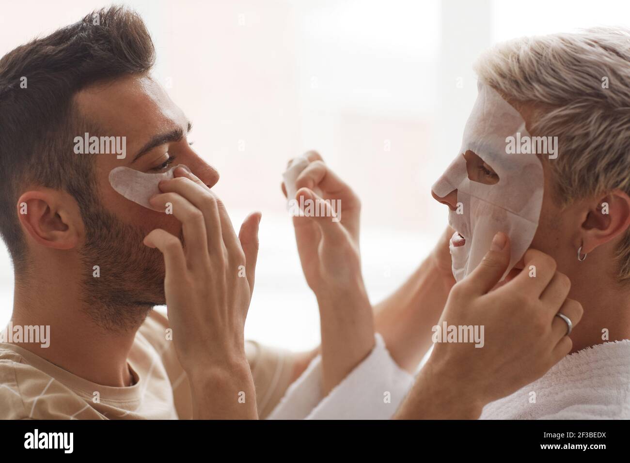 Vue latérale gros plan d'un jeune couple gay appliquant le visage masques les uns sur les autres tout en appréciant la routine de beauté du matin Banque D'Images