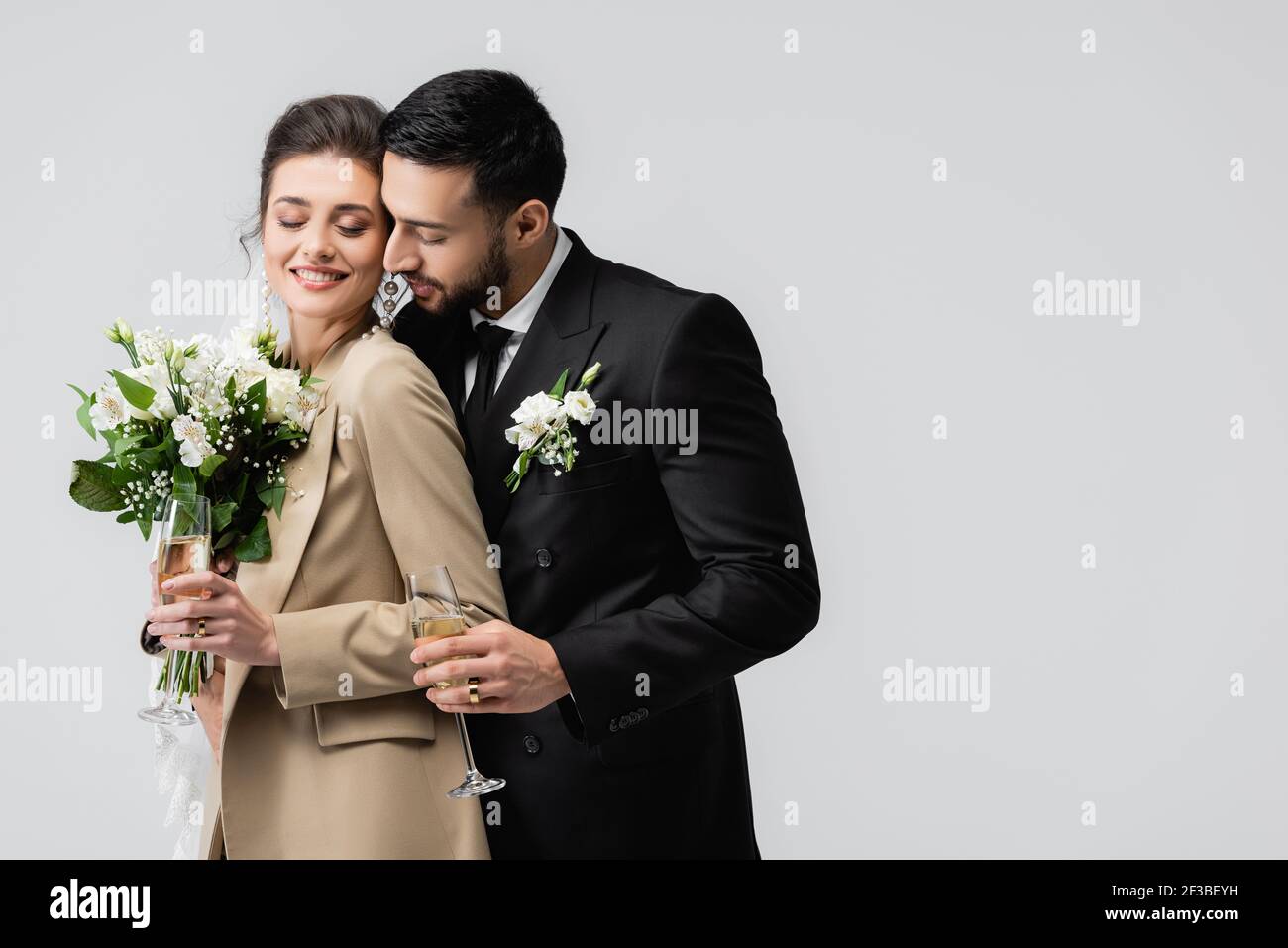 elégant homme arabe avec verre à champagne embrassant les mariés heureux tenue bouquet de mariage isolé sur gris Banque D'Images