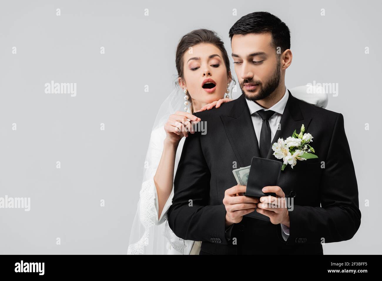 Mariée enthousiaste regardant le portefeuille avec l'argent entre les mains de marié musulman isolé sur gris Banque D'Images