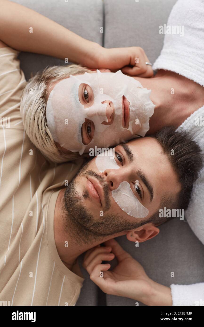 Vue de dessus gros plan d'un jeune couple gay regardant la caméra tout en étant allongé sur un canapé ensemble et portant des masques faciaux, beauté et soin de la peau concept Banque D'Images
