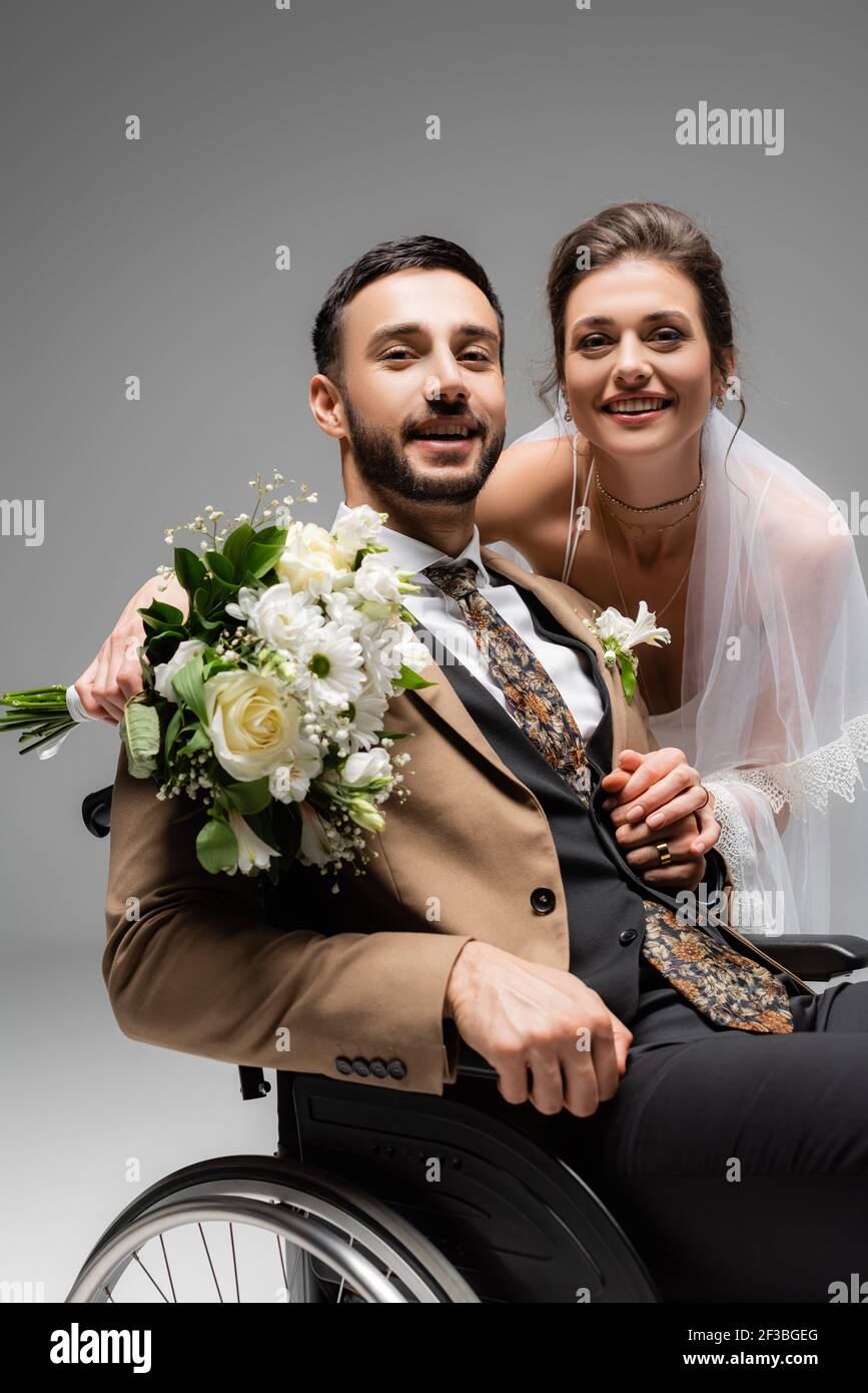 Marié arabe assis en fauteuil roulant près d'une mariée souriante isolée gris Banque D'Images