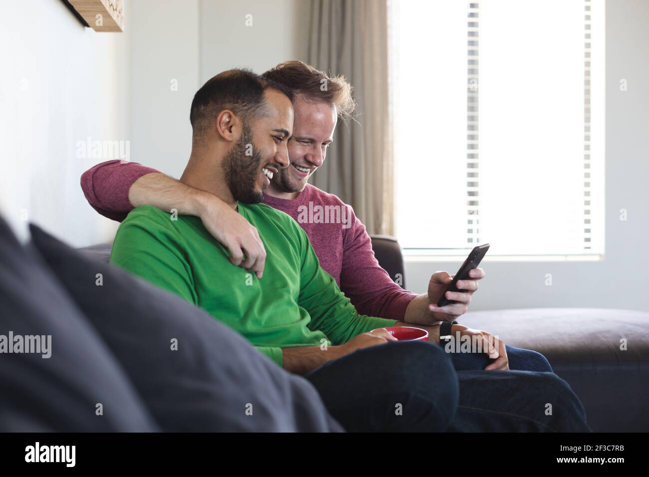 Couple multi-ethnique gay masculin souriant, assis sur un canapé et utilisant un smartphone à la maison Banque D'Images