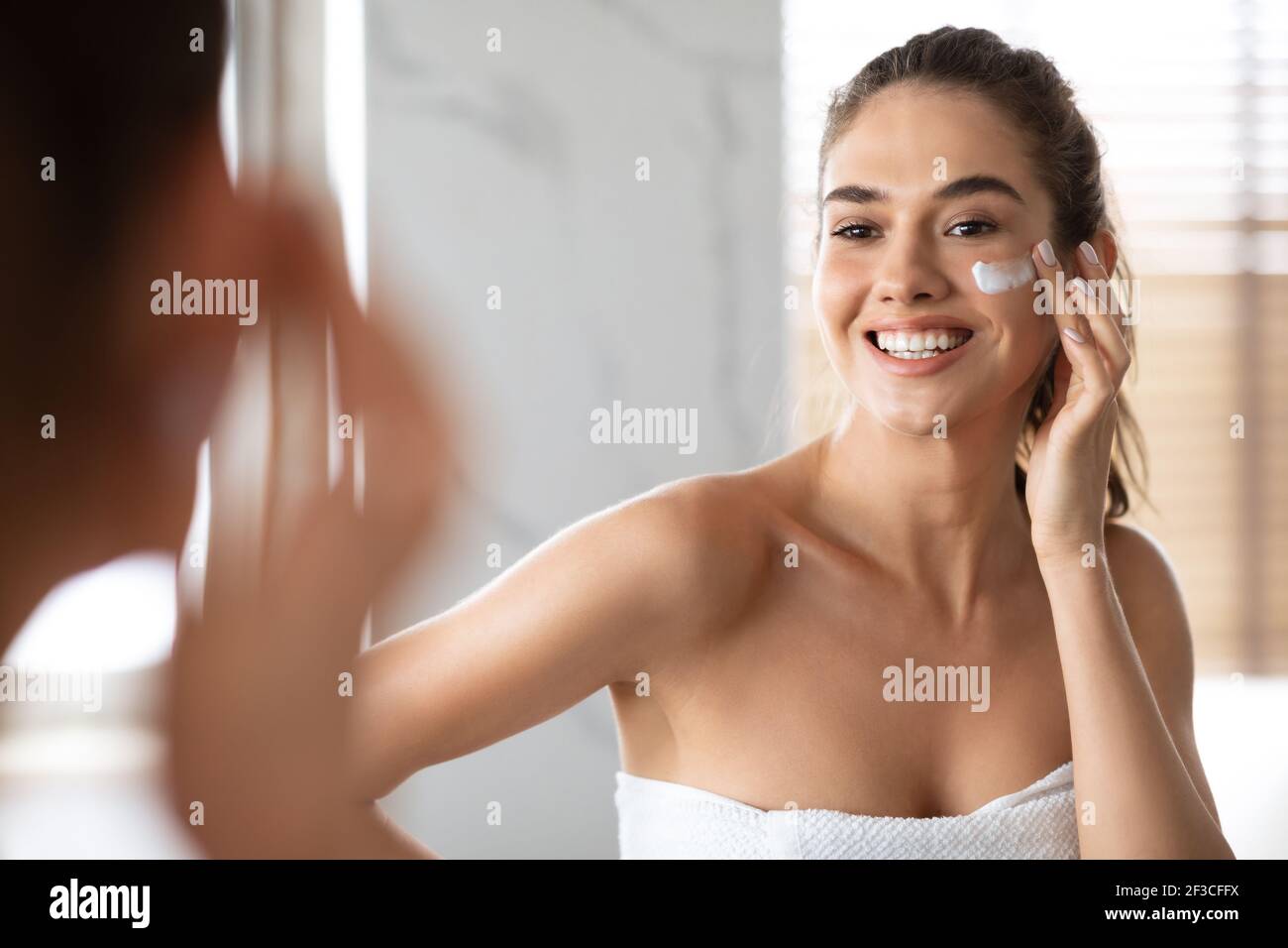 Femme appliquant de la crème sous les yeux peau hydratante posant dans la salle de bains Banque D'Images