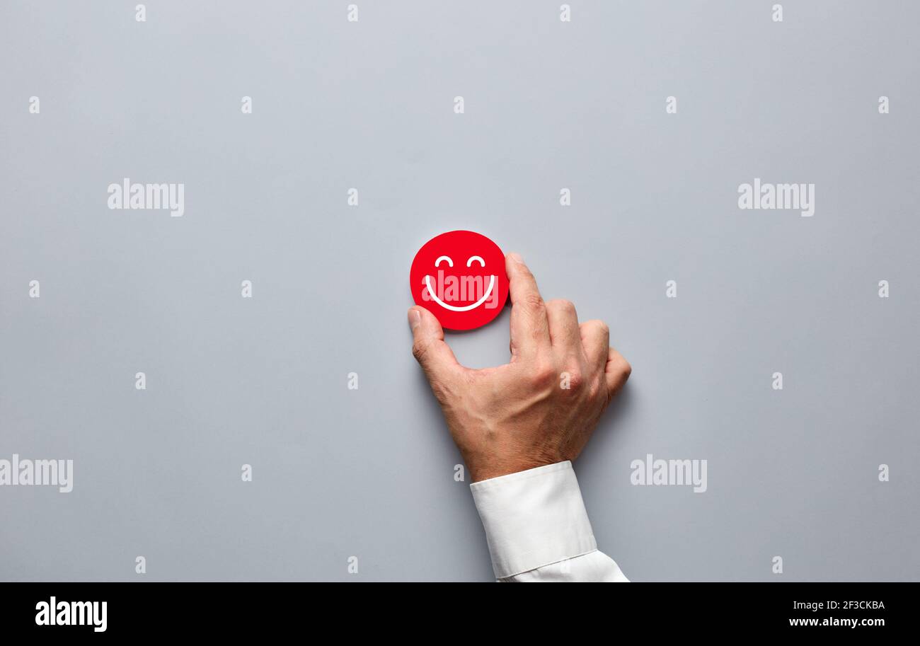 Main d'homme d'affaires tenant un badge rouge avec une icône de visage souriant. Satisfaction du client ou concept de feedback positif. Banque D'Images