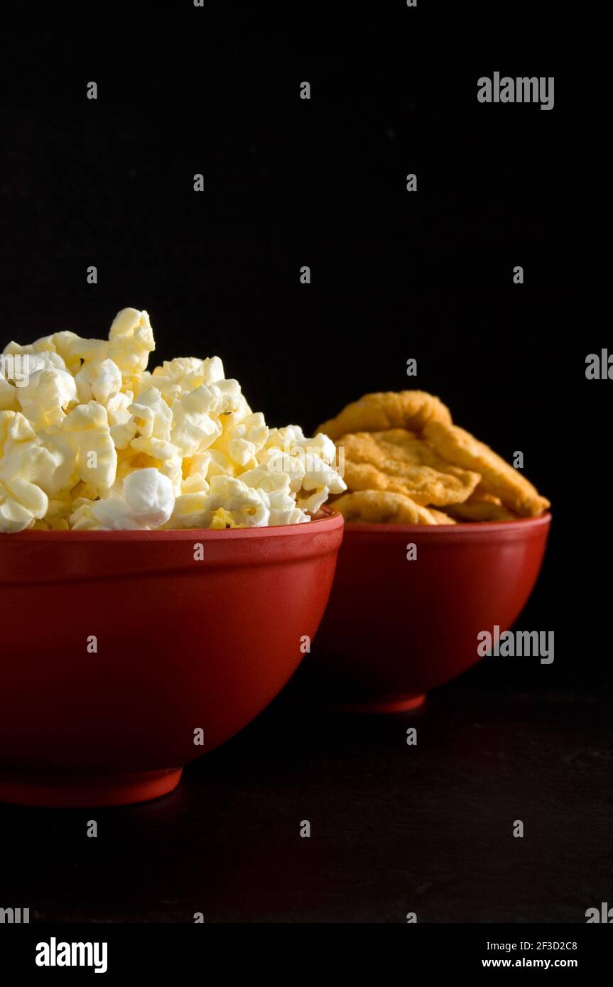 Vue latérale sur le popcorn beurré et les chips de riz au barbecue Dans les bols rouges isolés sur la verticale noire Banque D'Images