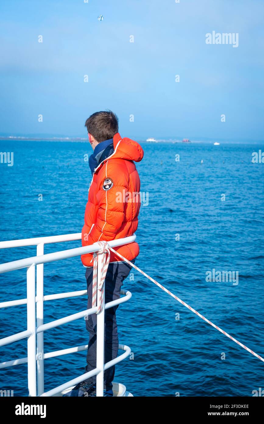 Homme dans des vêtements chauds debout sur la pointe avant du bateau, s'inclinant sur les rails et regardant loin. Journée ensoleillée en hiver sur la mer. Banque D'Images