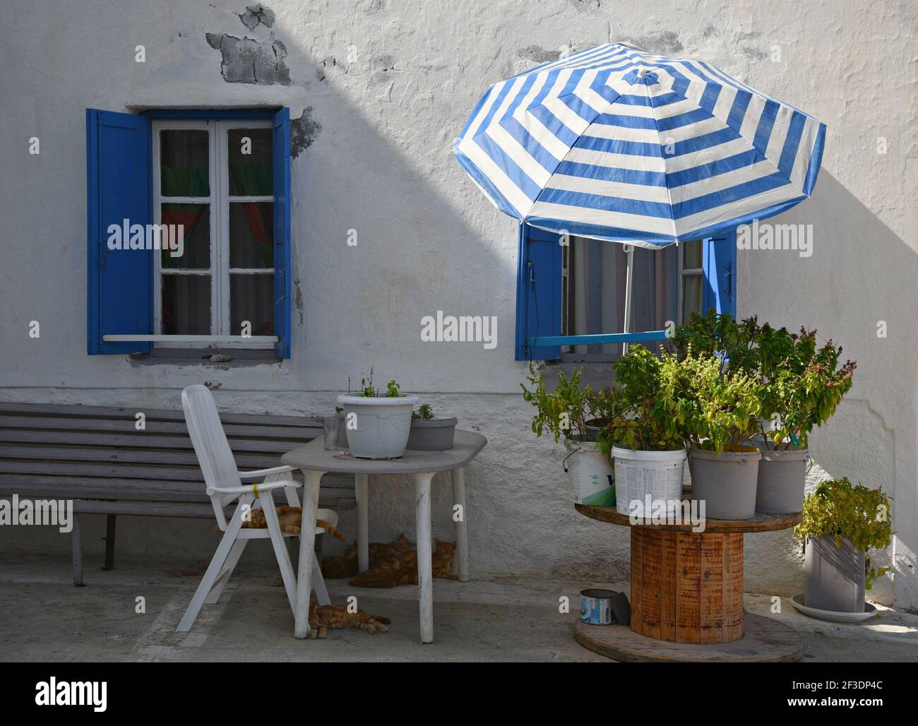 Façade traditionnelle de maison ancienne avec un mur blanc aux intempéries,  des volets en bois bleu, des meubles de patio et un parasol bleu et blanc  sur l'île d'Amorgos en Grèce Photo