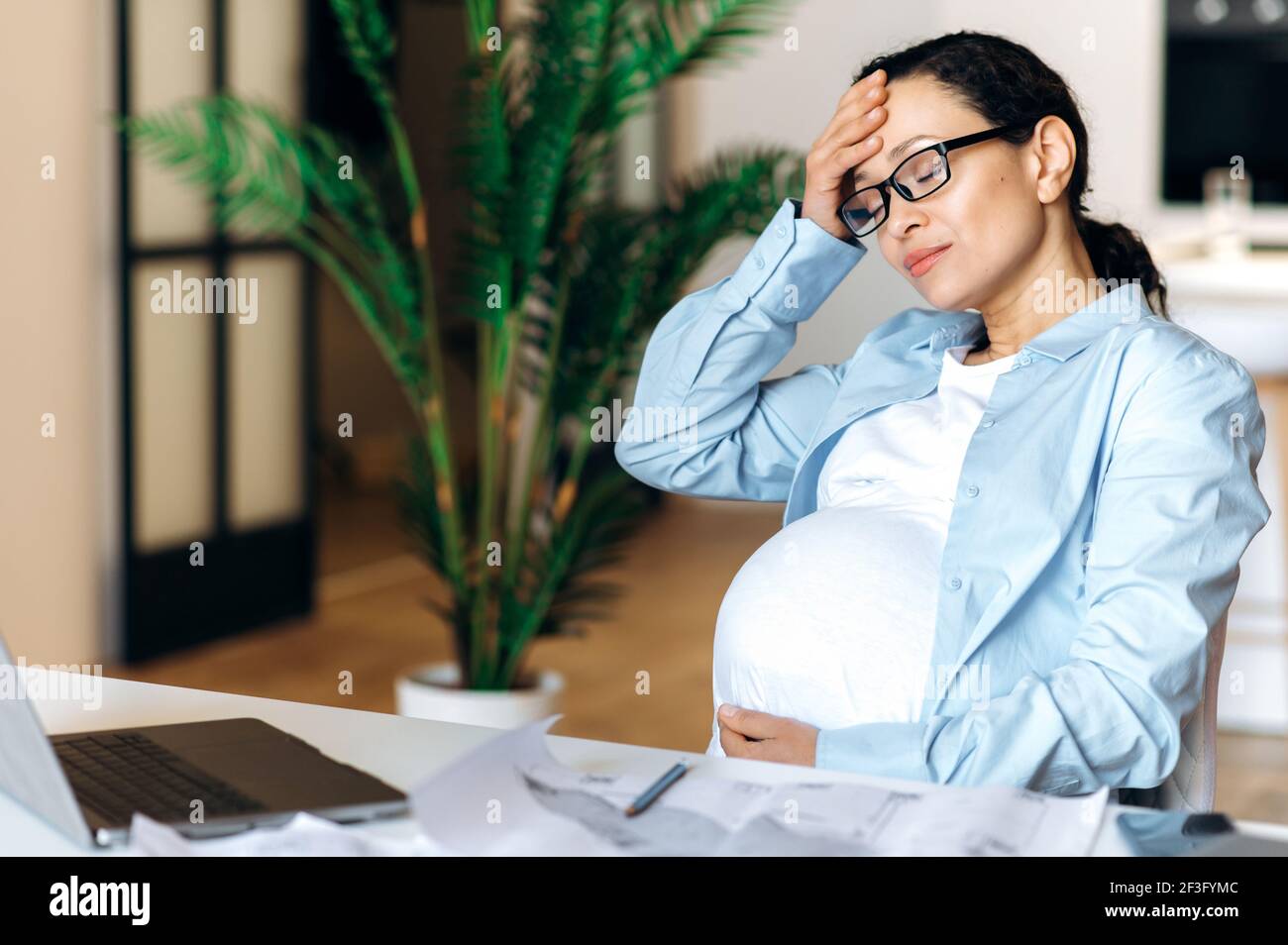 Exténuée femme enceinte mixte de race adulte assis à son bureau se sent un mal de tête, est stressé, tient sa tête avec sa main, les yeux fermés, fatigué de travail sédentaire avec ordinateur portable, besoin d'une pause et de repos Banque D'Images