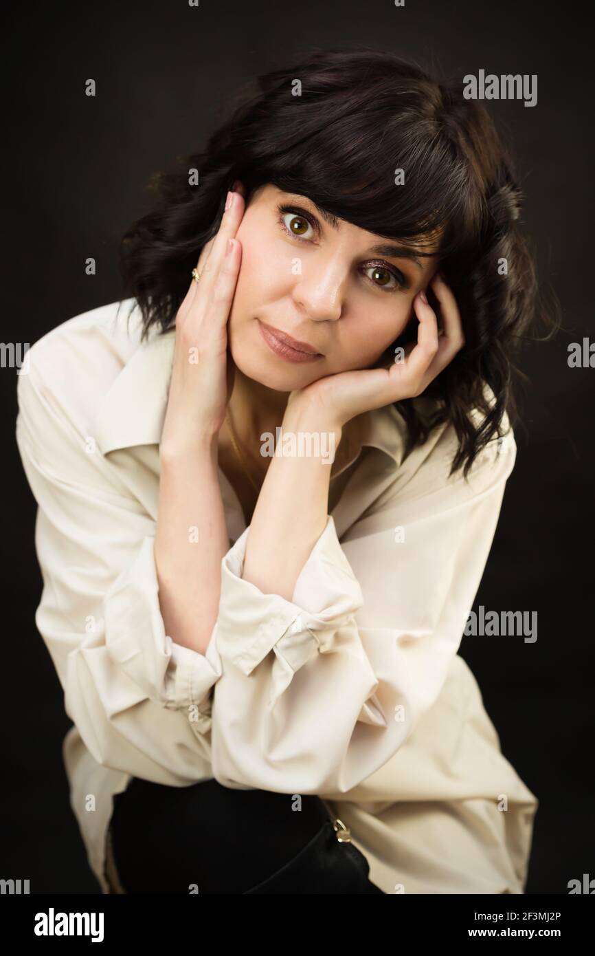 Brunette avec les yeux bruns dans une chemise blanche embrassait son visage avec ses mains sur un fond noir. Banque D'Images