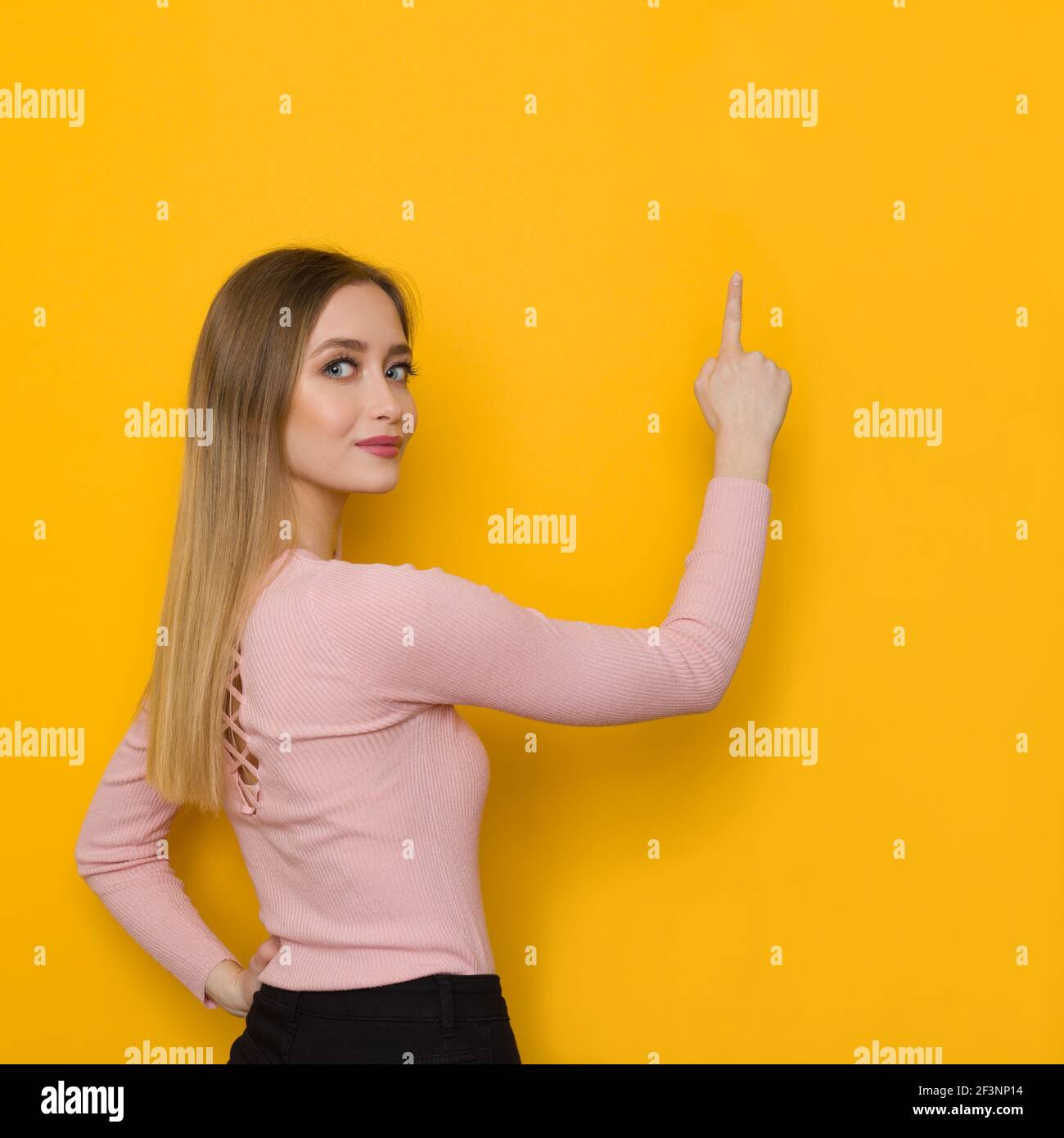 La jeune femme en chandail rose pointe vers le mur jaune et regarde l'appareil photo au-dessus de l'épaule. Vue arrière. Prise de vue en studio à taille haute. Banque D'Images
