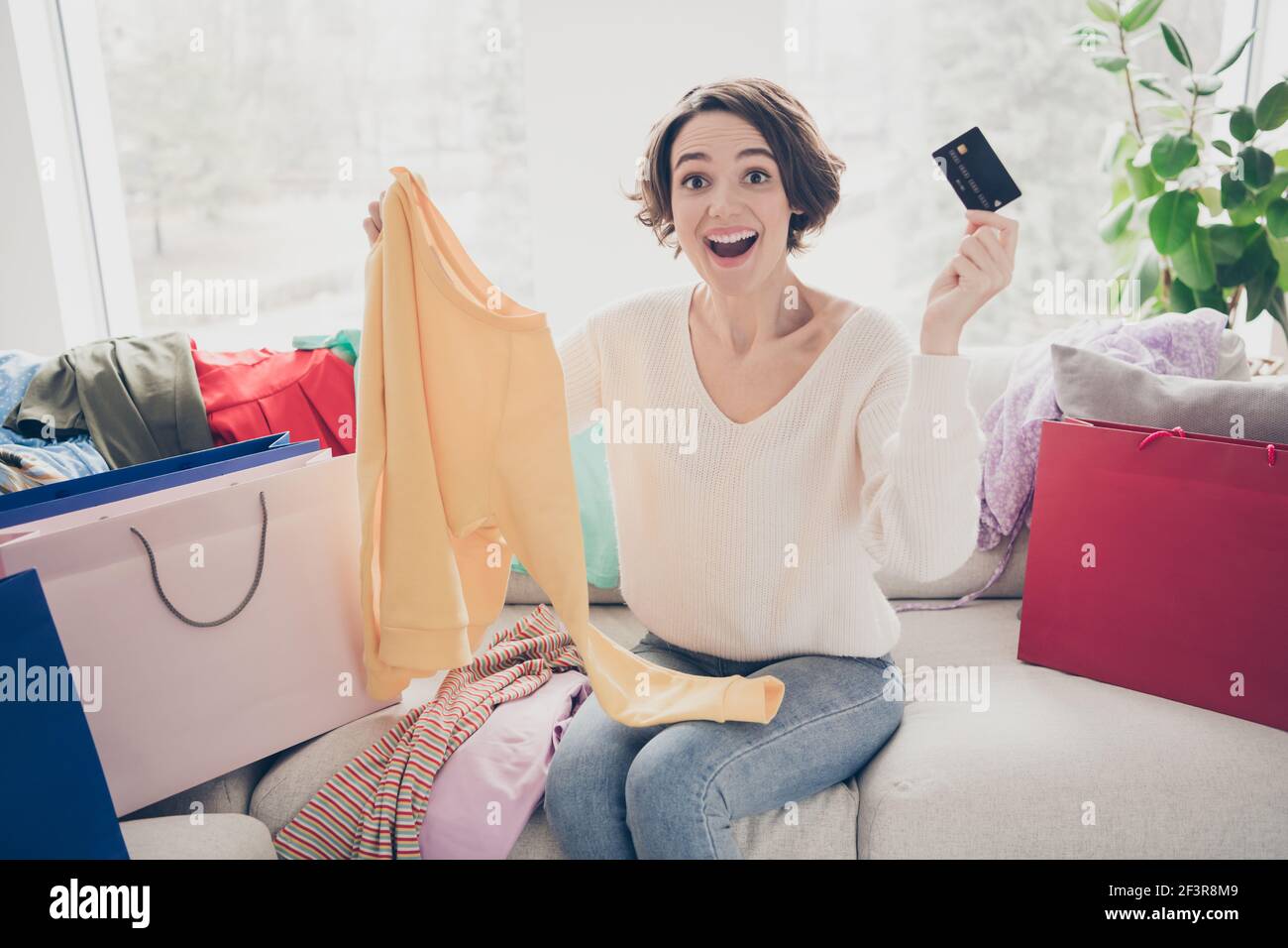 Photo de la jeune femme excitée choquée tenir le spectacle de carte de crédit chandail solde sacs à provisions tenir le chandail à l'intérieur de la maison Banque D'Images